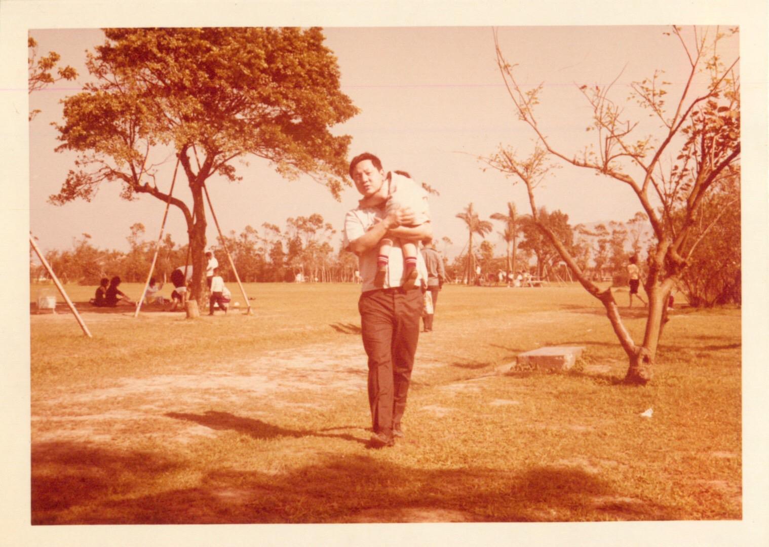 A father carries his child while walking in a park filled with families and greenery.