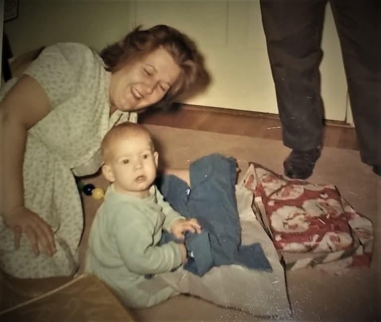 A happy toddler explores a wrapped present while an adult watches joyfully in a warm room.