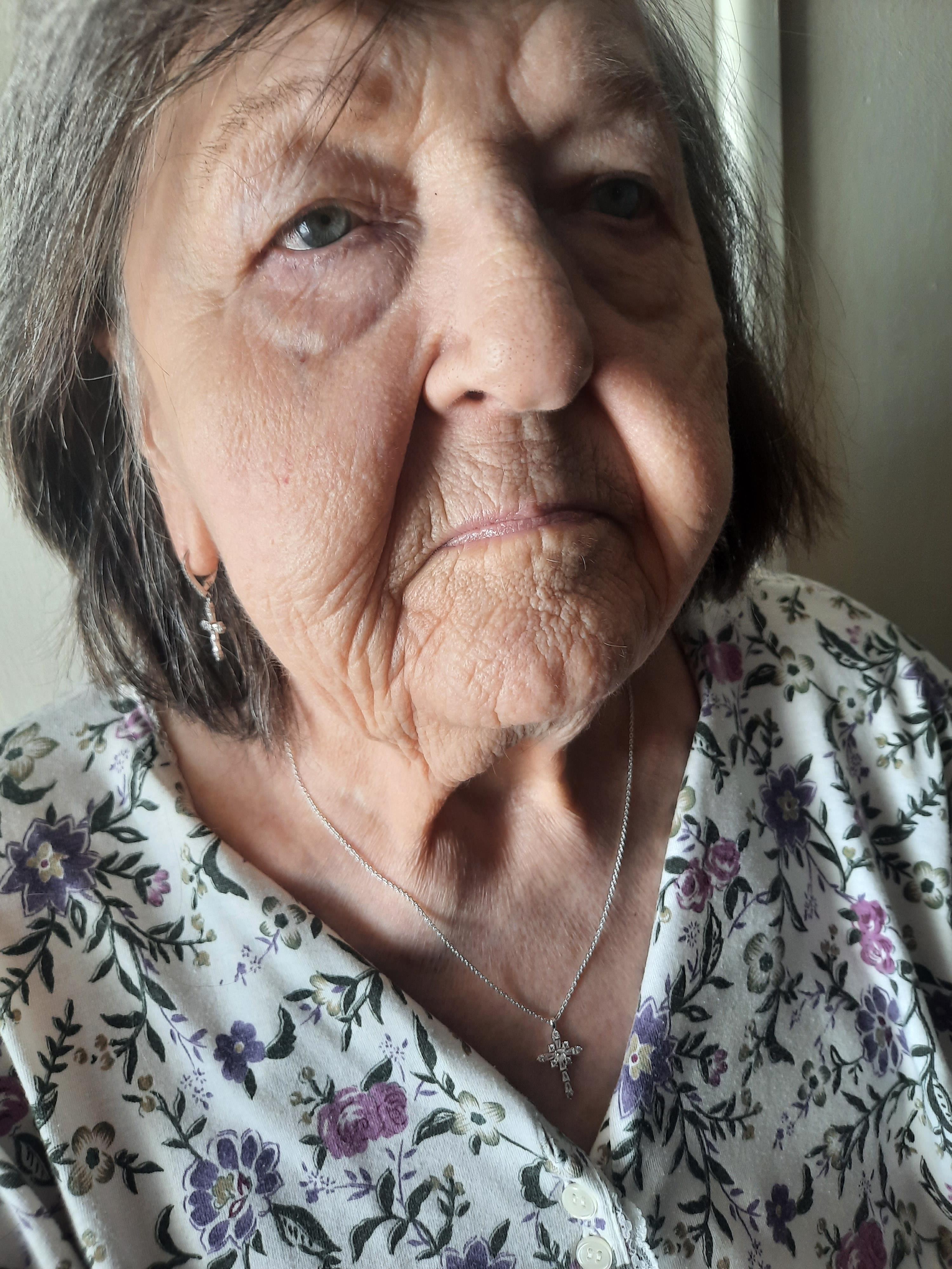An elderly woman sits indoors, calmly wearing floral clothes and delicate jewelry.