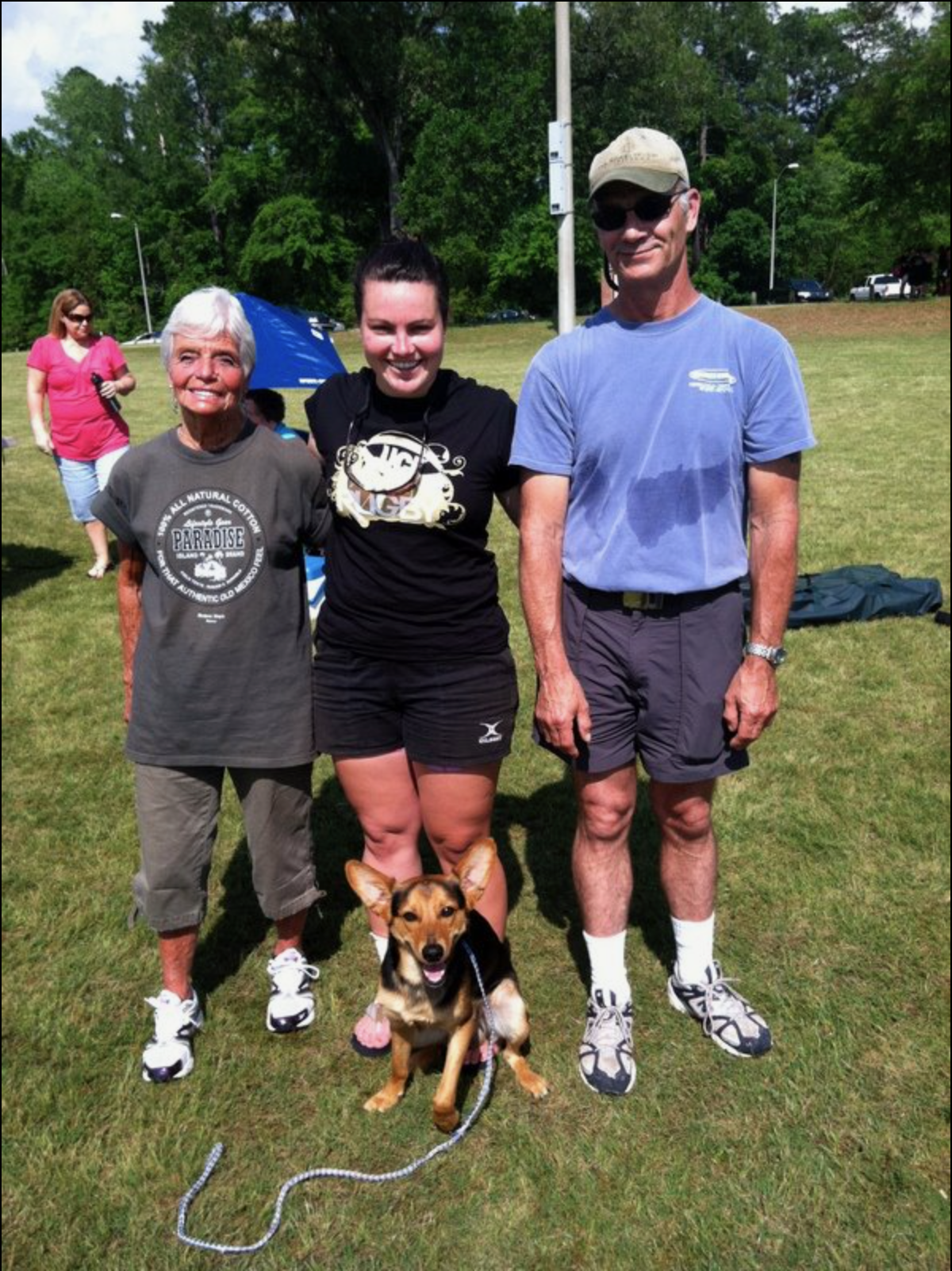 Three people pose with a dog at a park while others enjoy a sunny day outdoors.