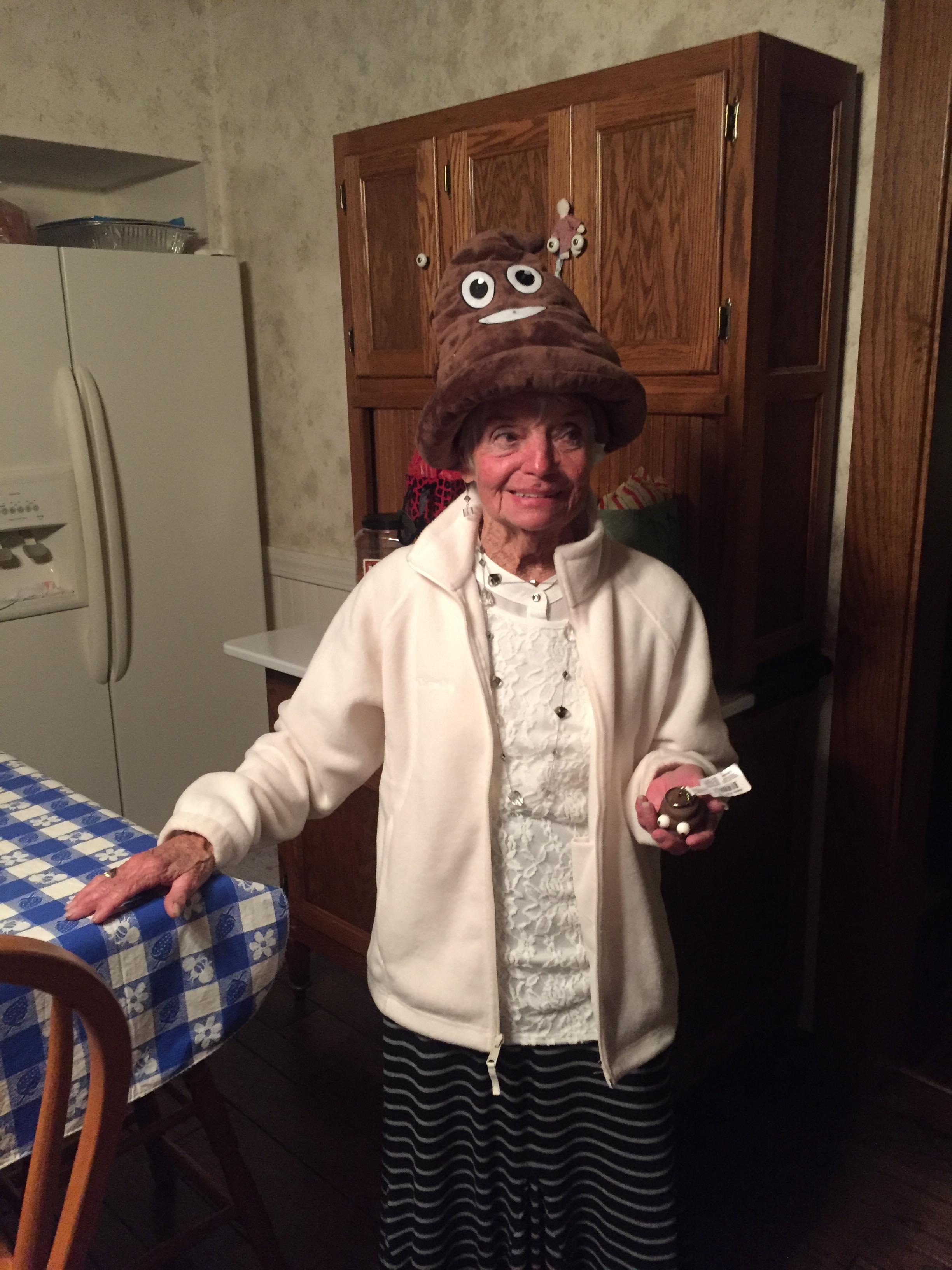 An elderly woman stands in a warm kitchen, wearing a comical hat and holding a small treat.