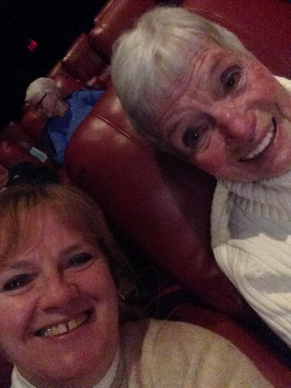 Two friends smile brightly while watching a movie in comfortable red seating at the theater.