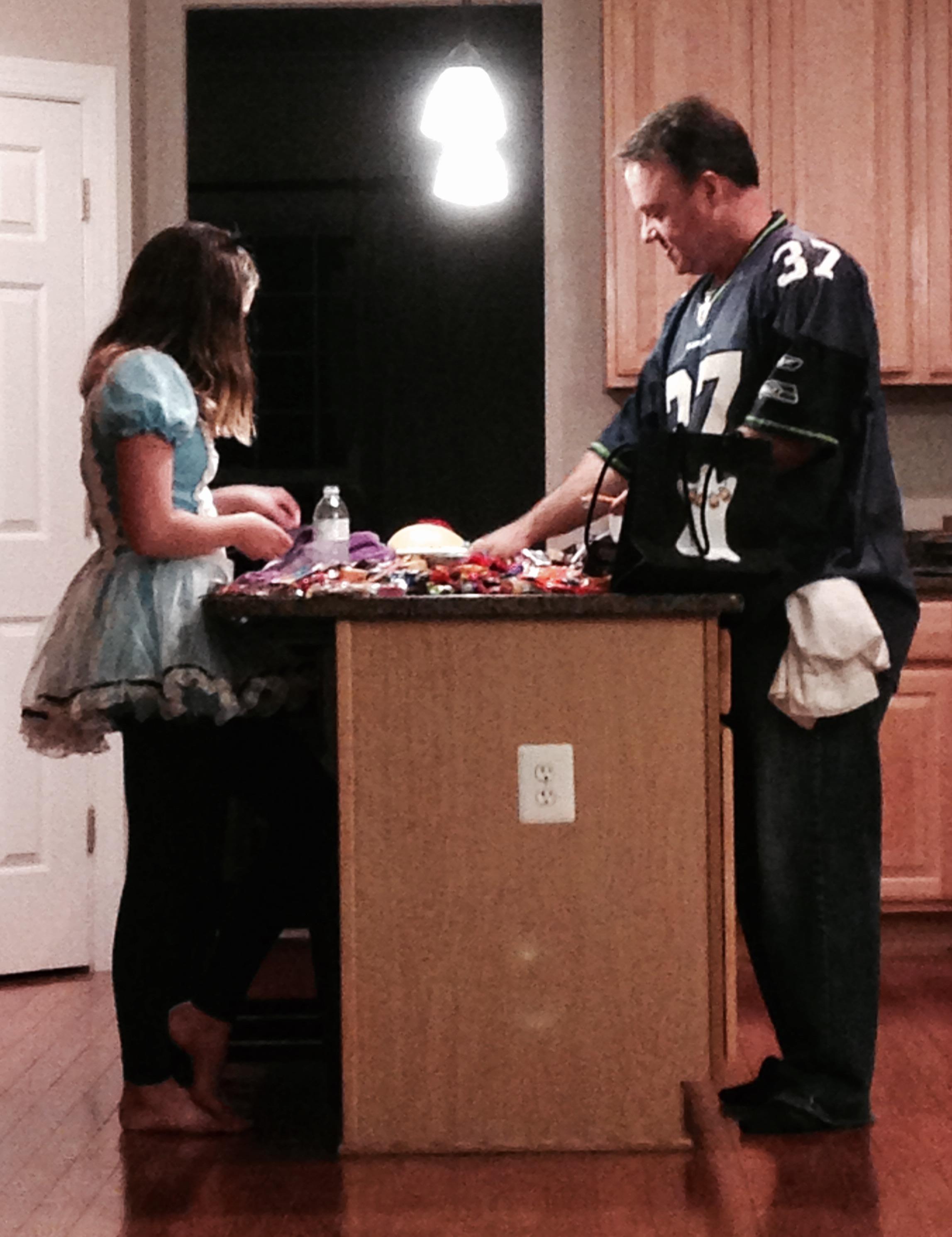 A girl in a costume and her father sort through candy and decorations for Halloween.