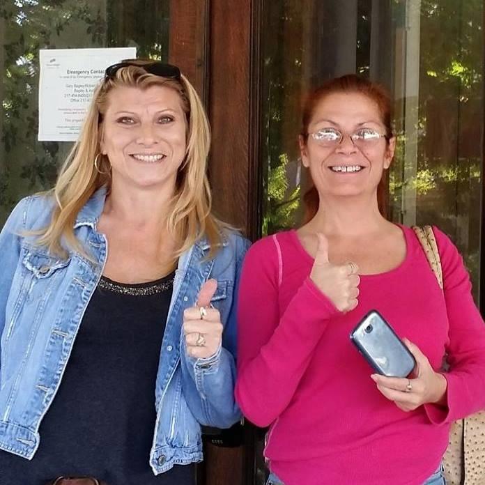 Two happy women stand outside a building, smiling and showing thumbs up, enjoying their time.