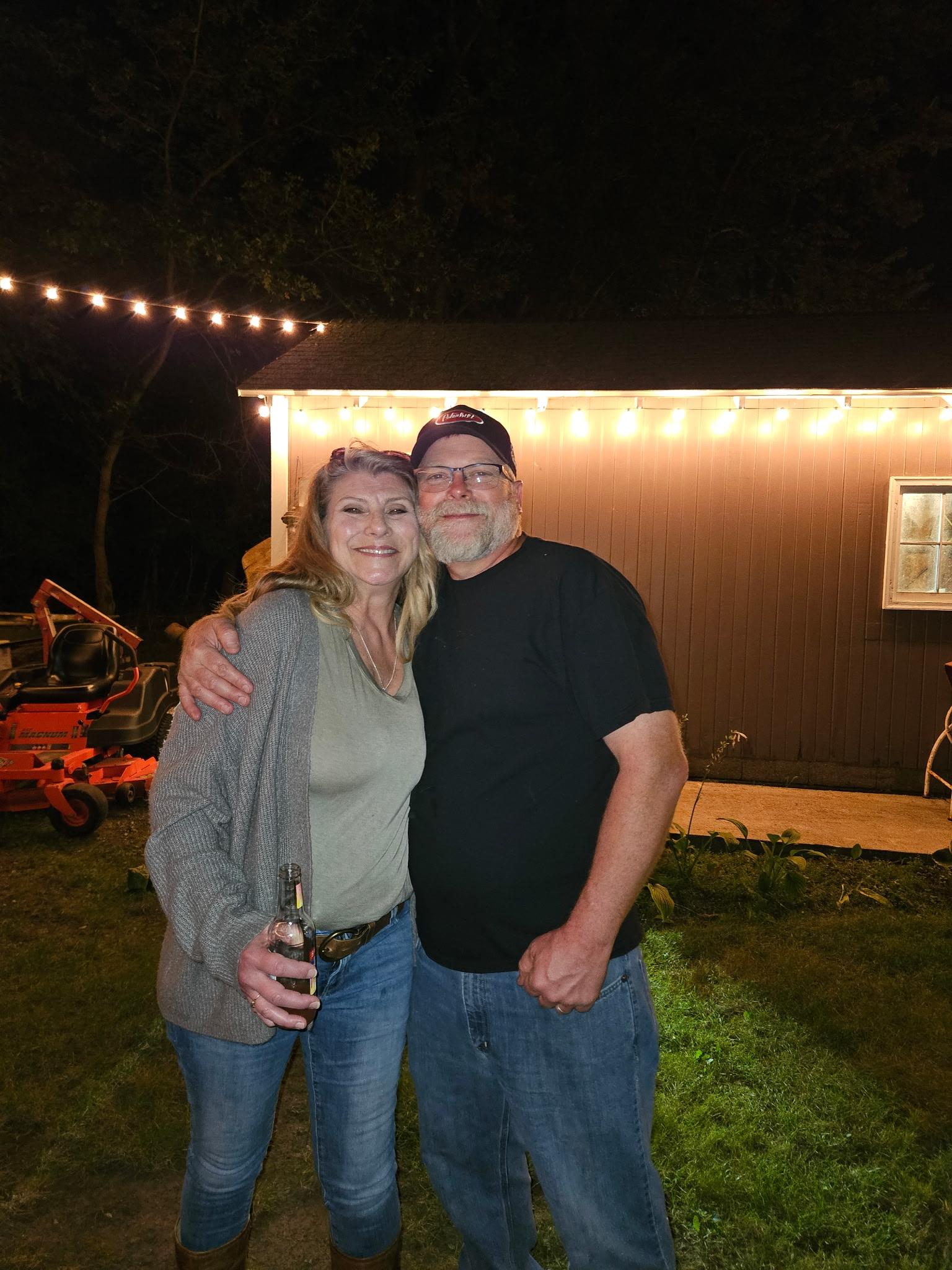 Two friends smile and embrace at a cozy backyard party illuminated by string lights.
