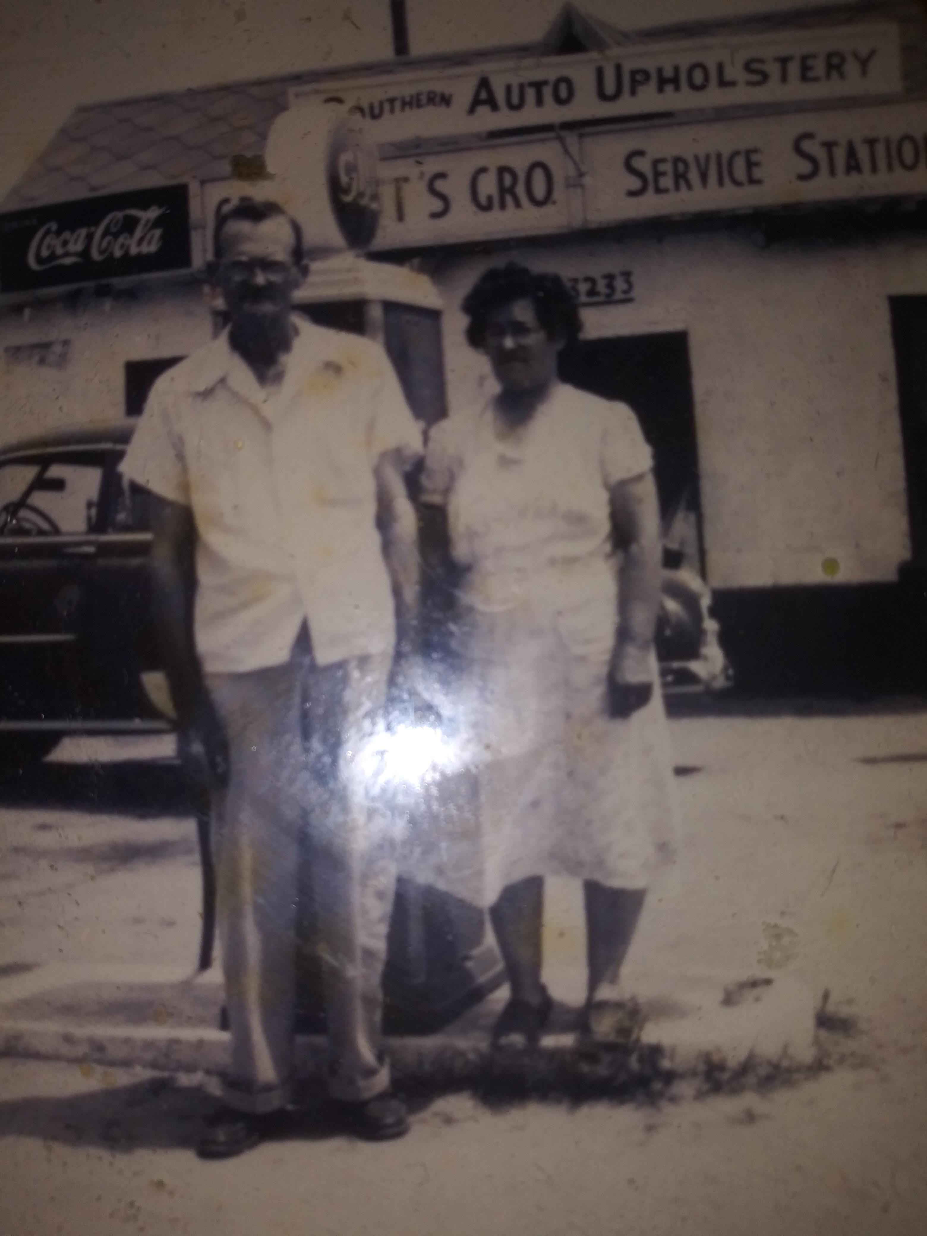 Couple poses together by a classic gas pump outside a service station during mid-20th century.