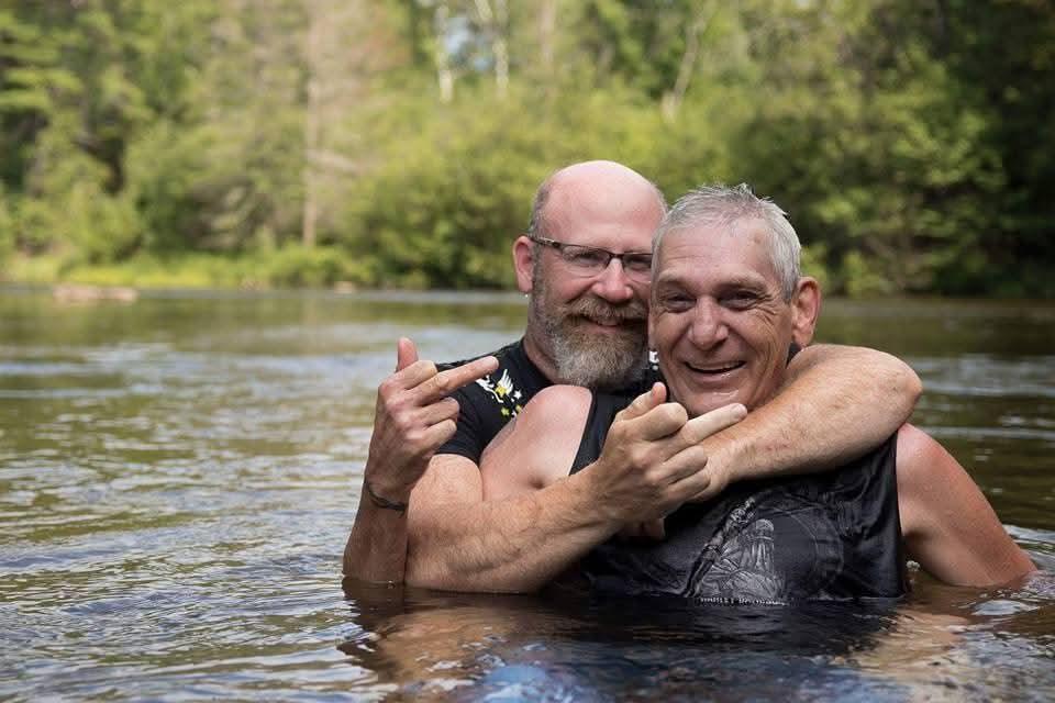 Two men share laughter and joy in the water, surrounded by lush greenery on a sunny day.