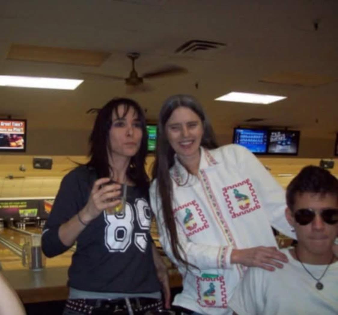 Two friends pose joyfully at a bowling alley while enjoying drinks and laughter.