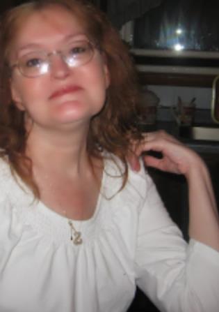 A woman with red hair and glasses is smiling happily, sitting in a kitchen at dusk.