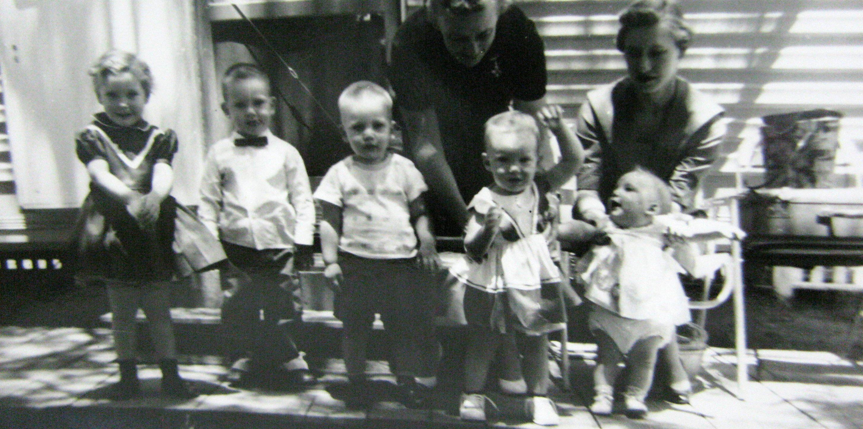 Six children and two adults stand together, smiling, in a backyard during the 1950s.
