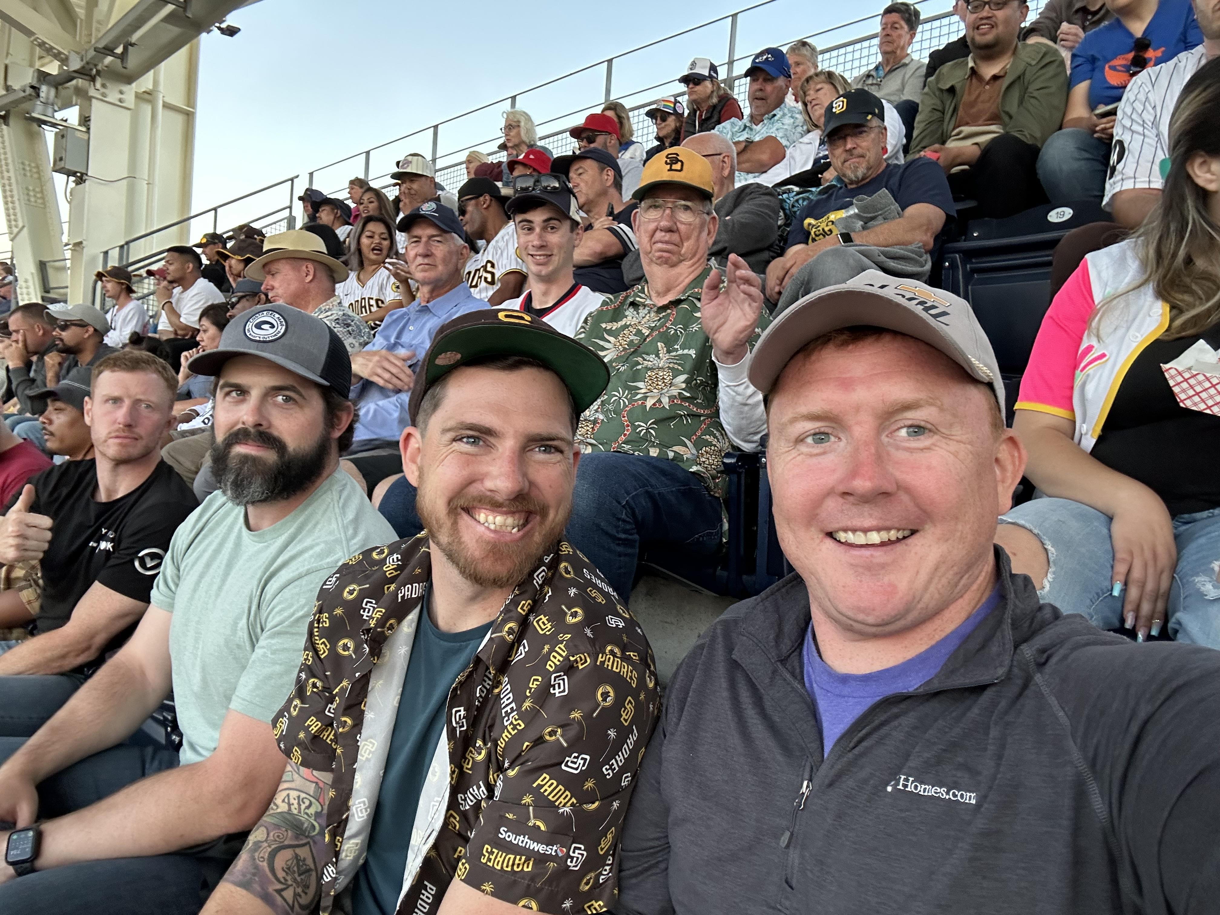 Group of friends smiles for a selfie at a baseball game in a crowded stadium on a sunny day.