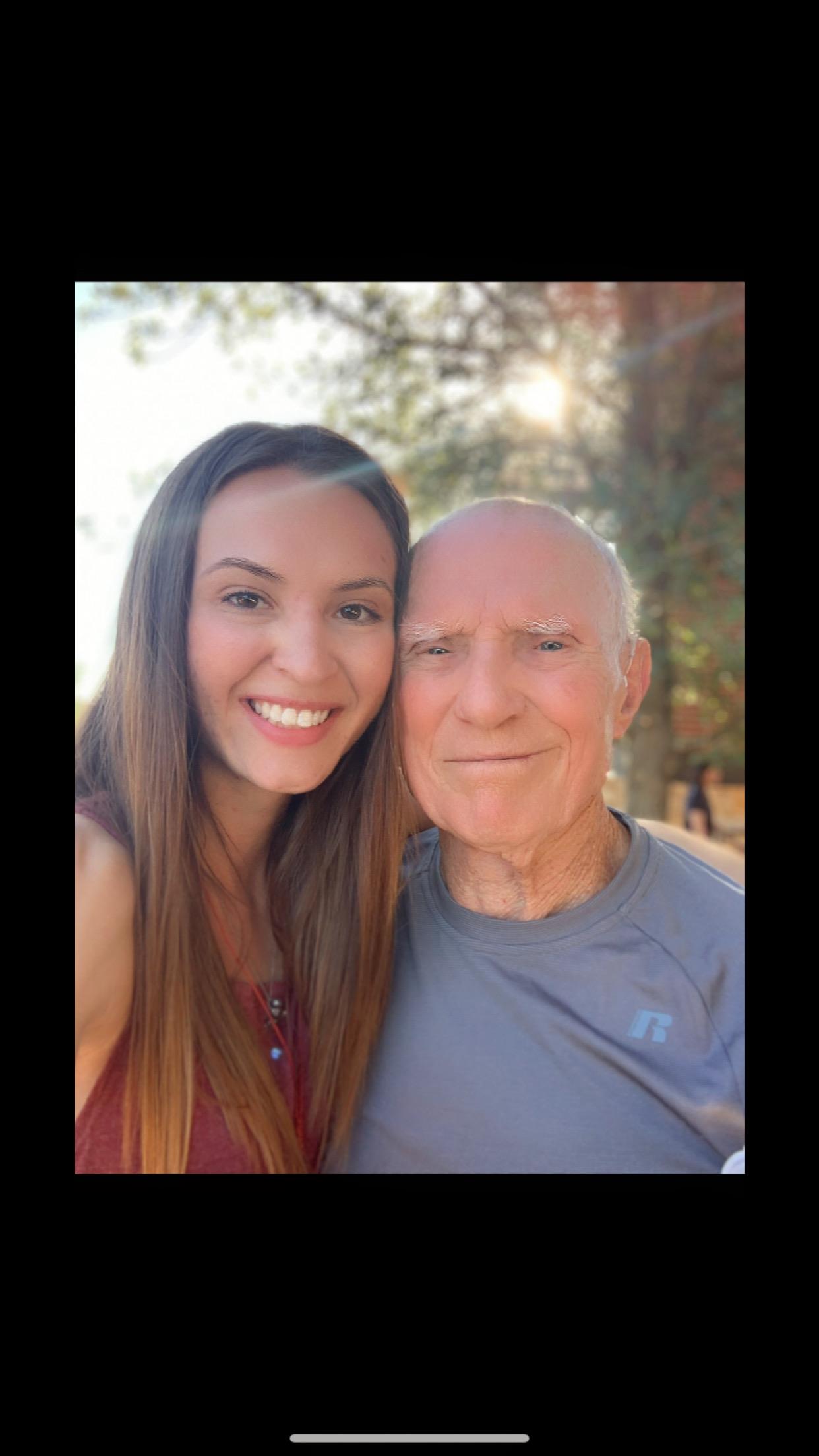 A young woman and a senior man pose together with joyful expressions in a park on a sunny day.