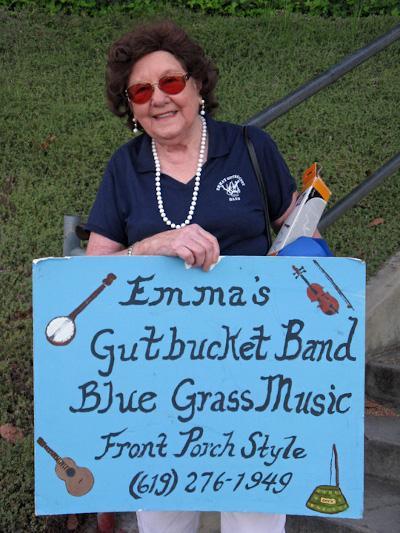 A smiling woman stands beside a colorful sign, promoting bluegrass music in a lively atmosphere.