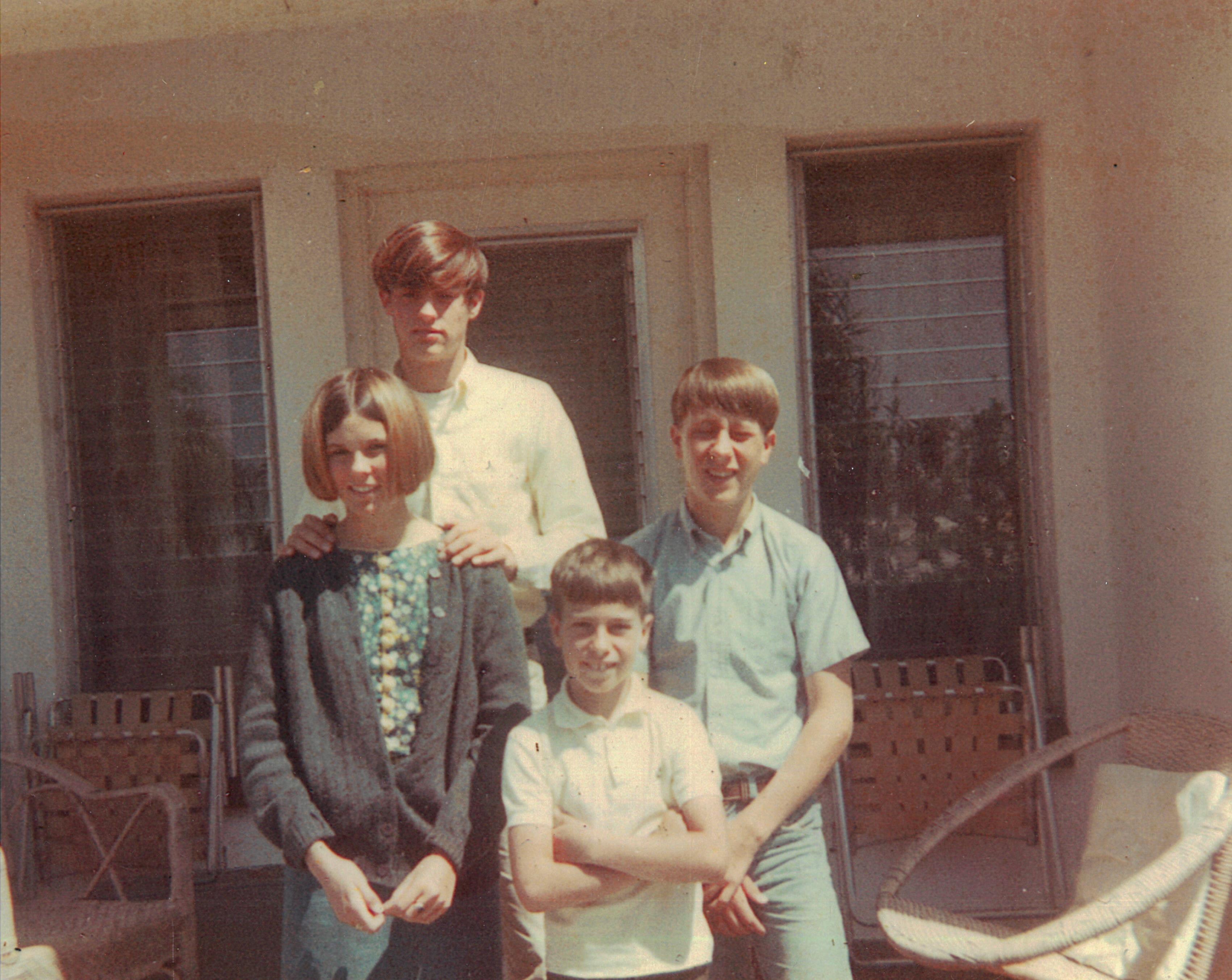 Four family members pose together on a porch, smiling in casual attire on a bright day.