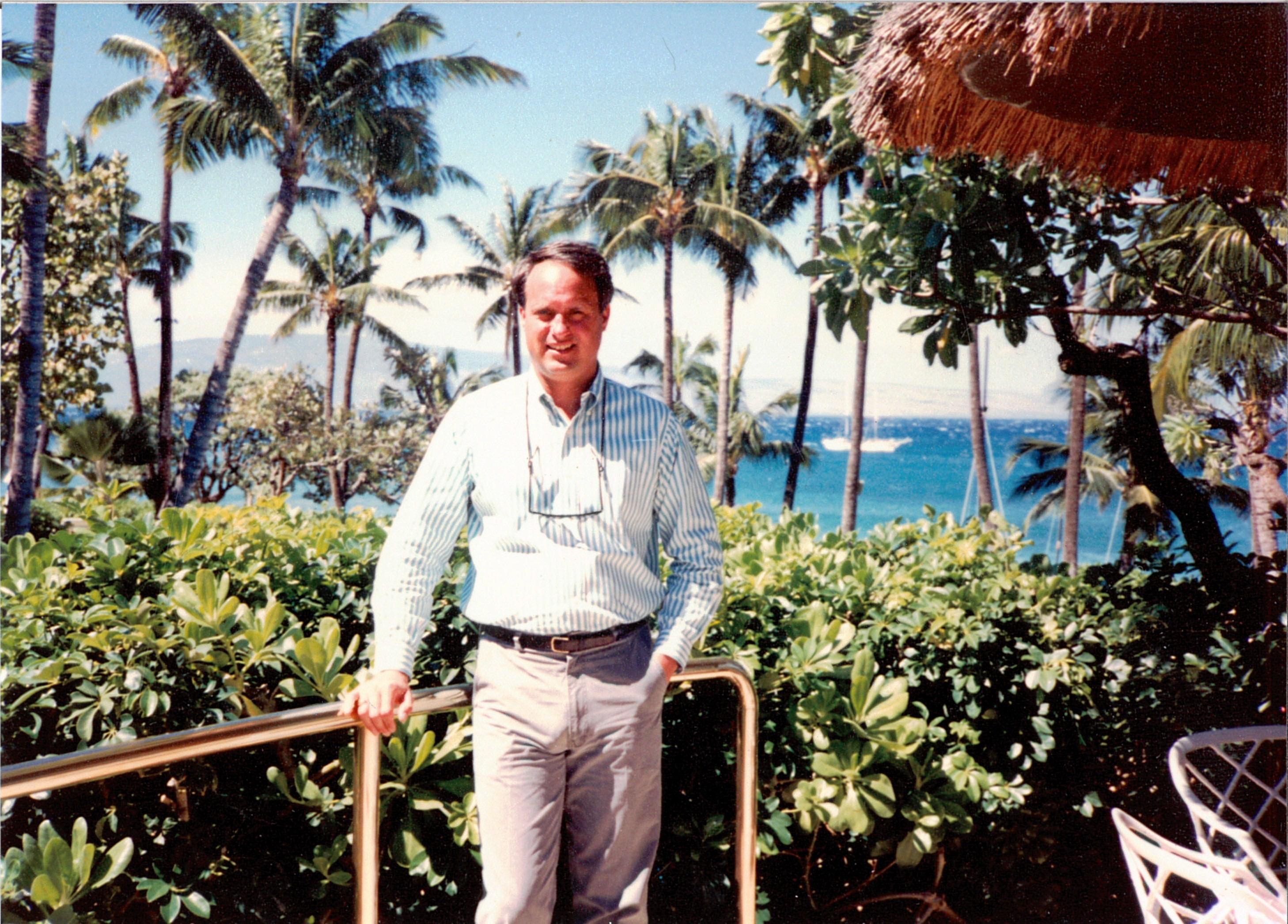 A man smiles by a railing, surrounded by tropical plants and ocean views under a clear sky.