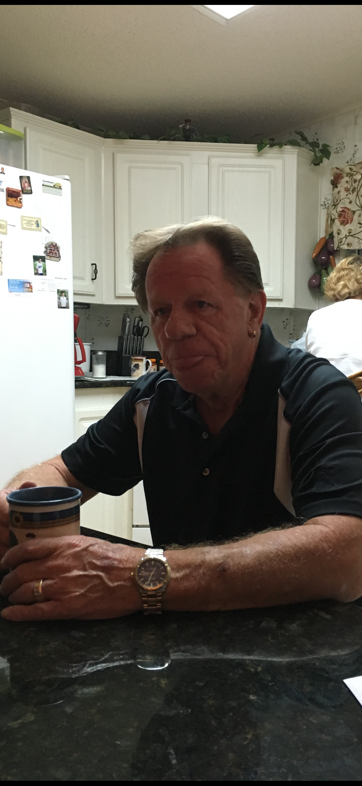 A man relaxes at a kitchen table with a cup of coffee as someone prepares food.