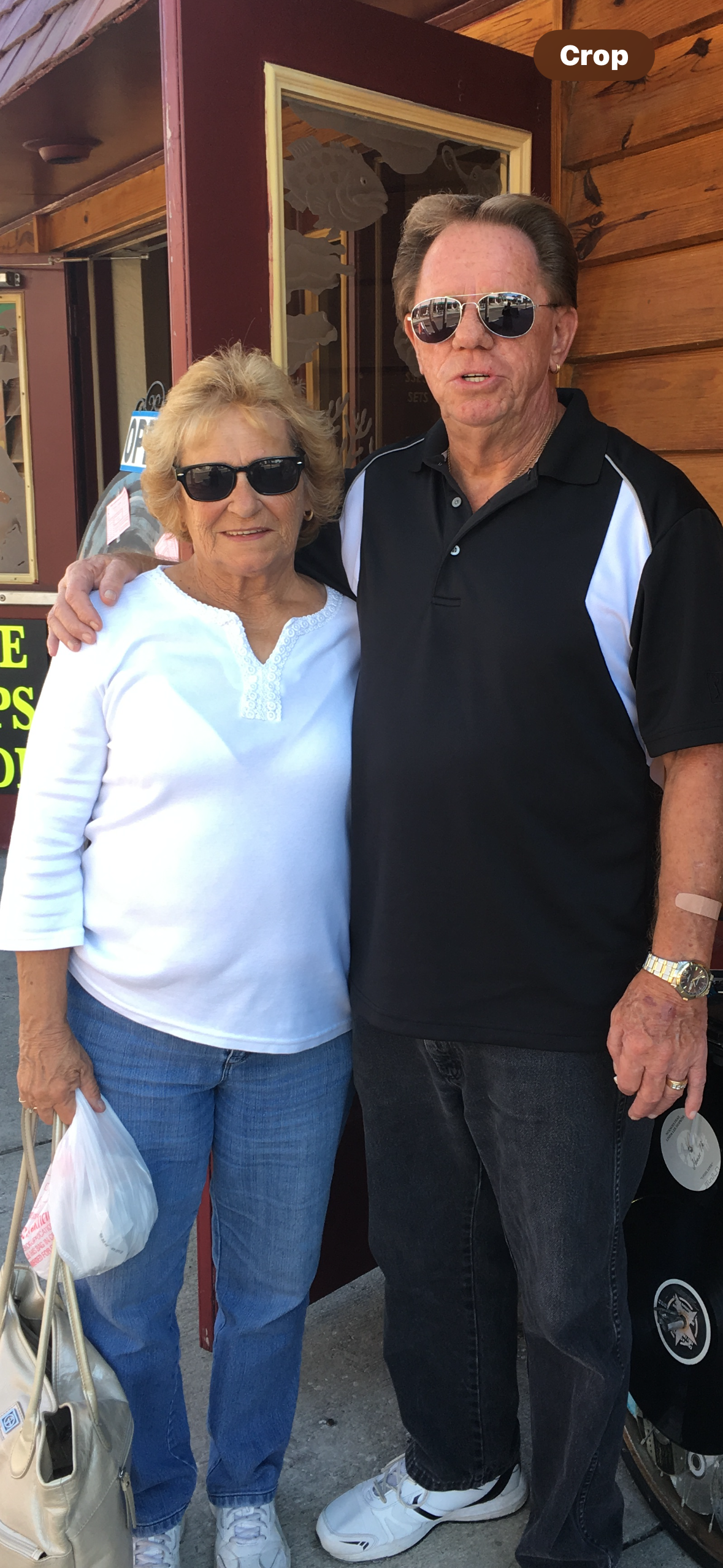 Elderly couple smiles while standing together outside a restaurant on a bright day.