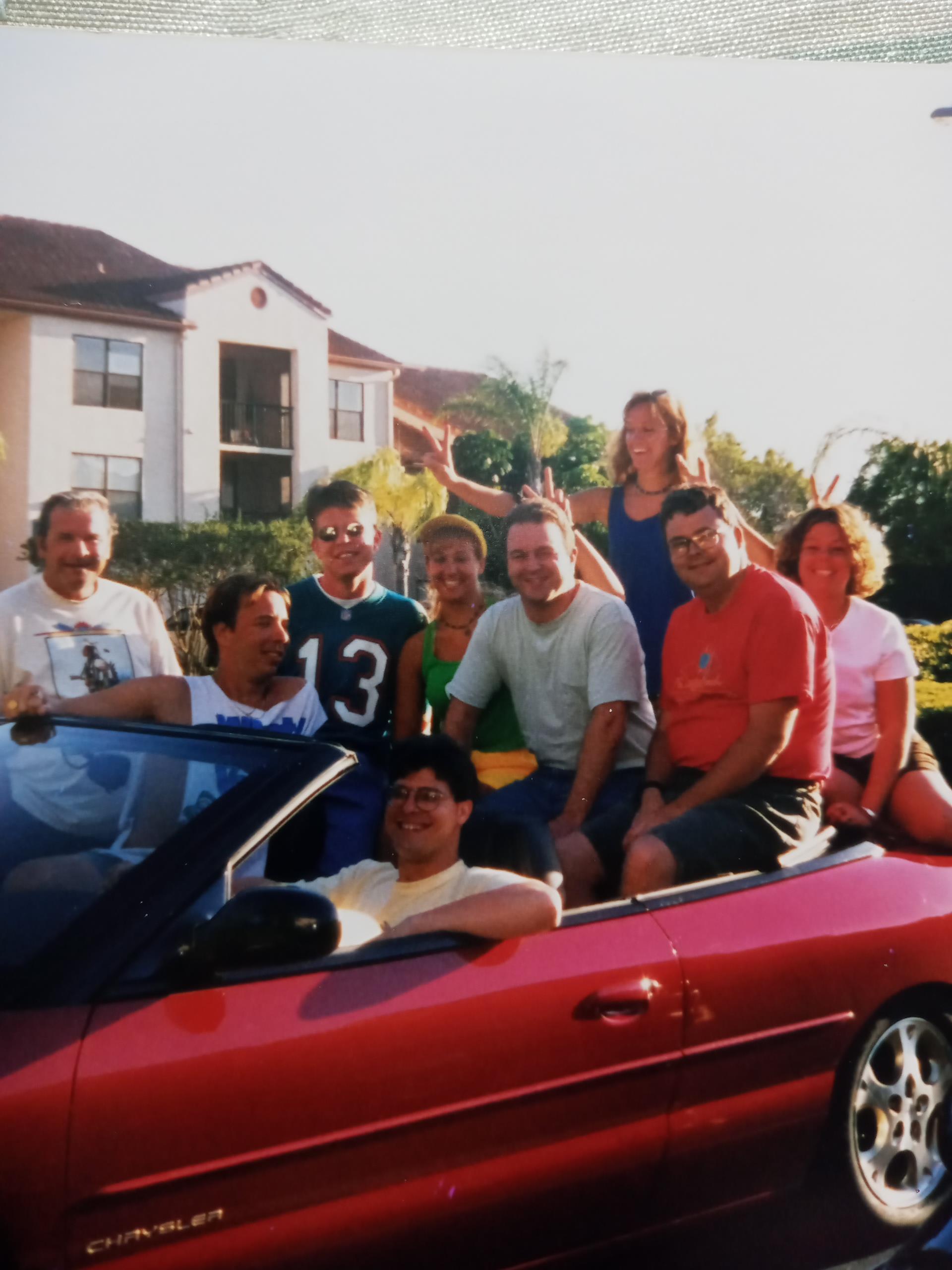 A group of friends gathered in a red convertible, soaking up the sun outside a suburban home.