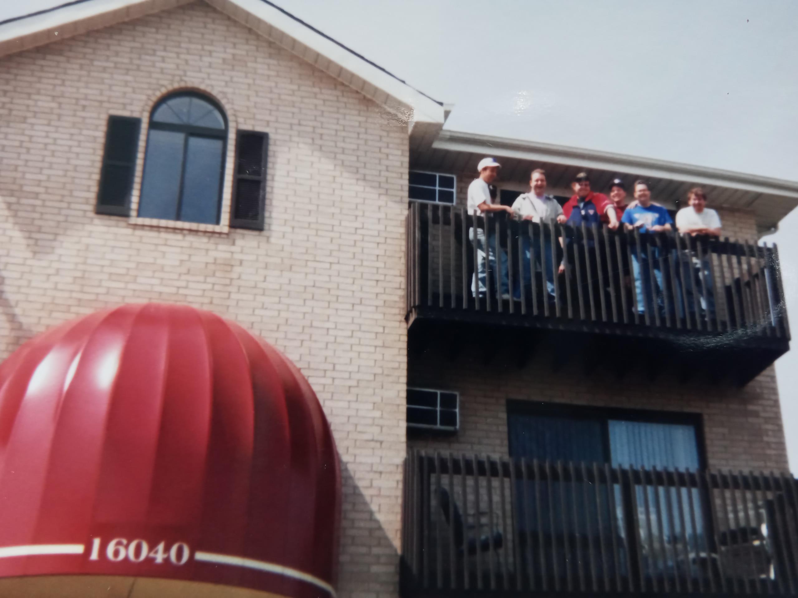 Friends smile and pose on balcony of a brick building on a bright, sunny day.