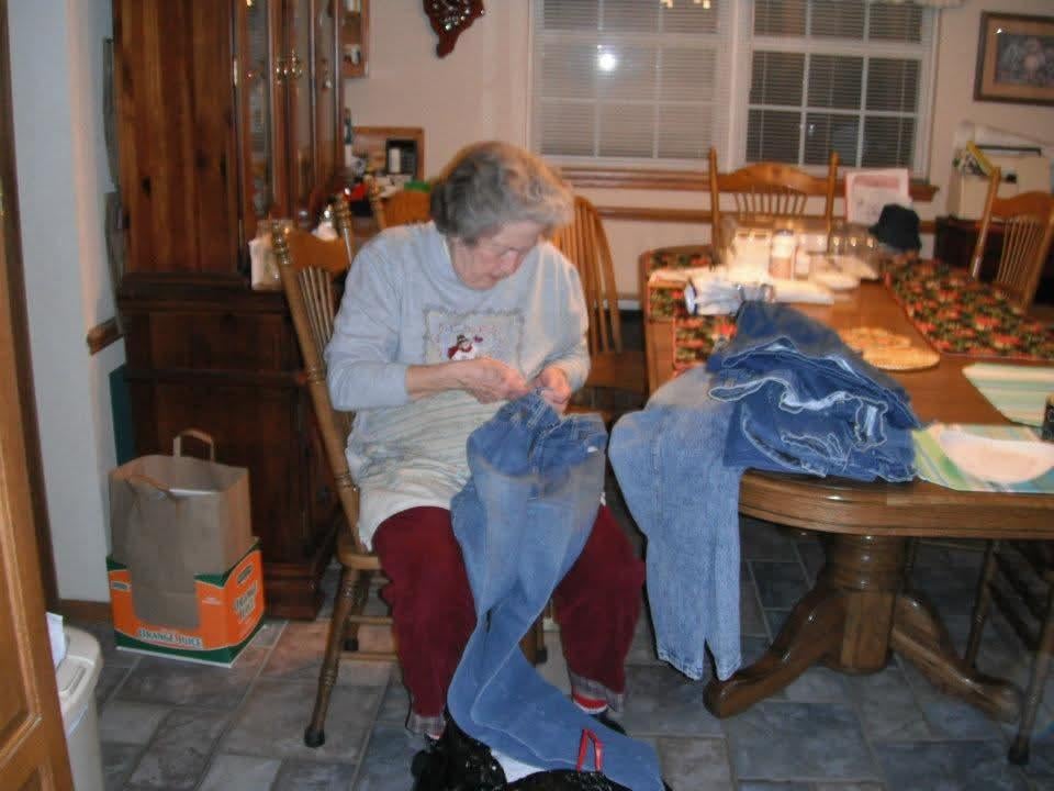 A woman works on sewing denim jeans at home, surrounded by wooden furniture and soft lighting.