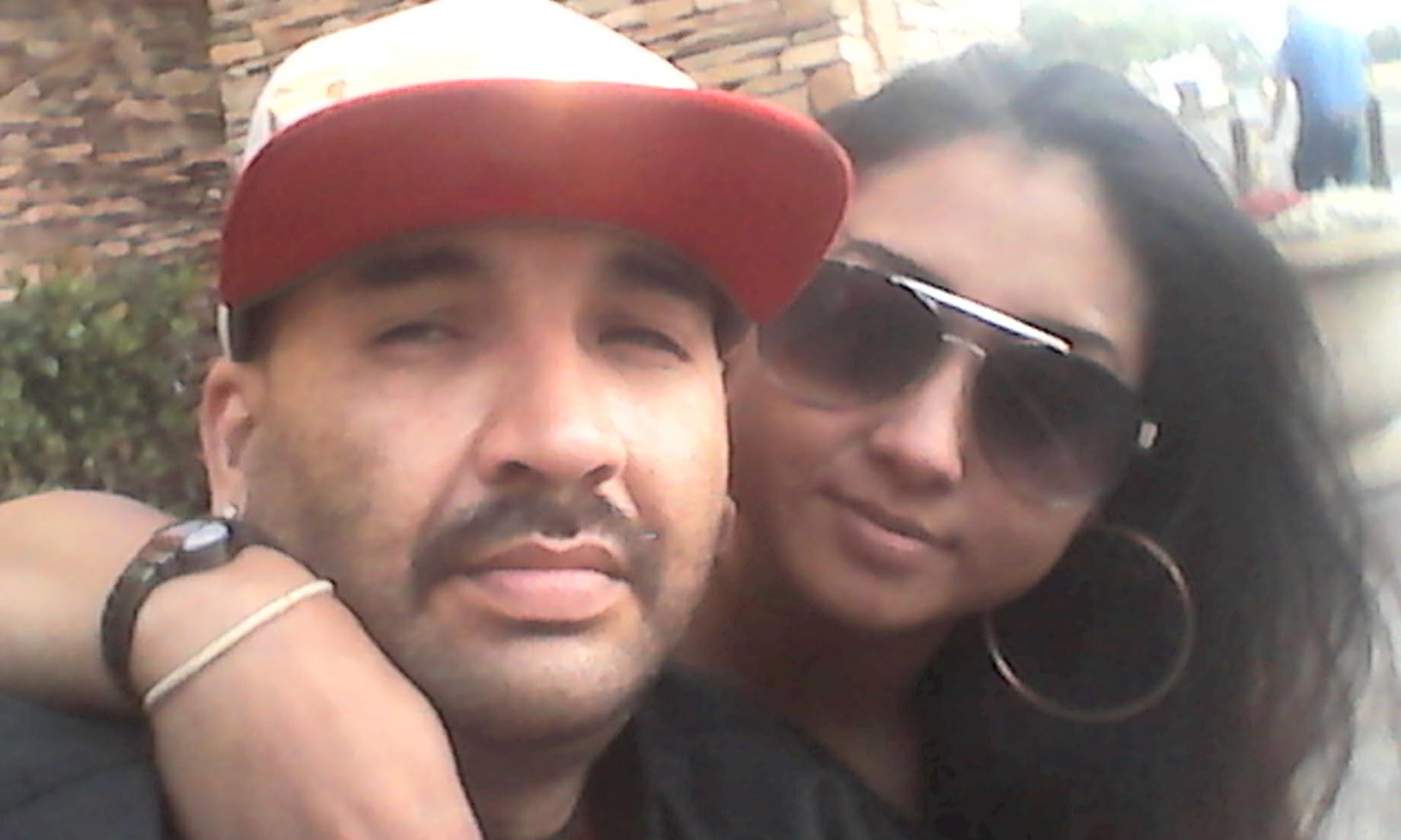 Two individuals pose closely together, smiling, in a lively outdoor setting under bright sunlight.