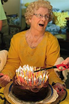 An elderly woman in a yellow outfit joyfully prepares to blow out candles on a birthday cake.