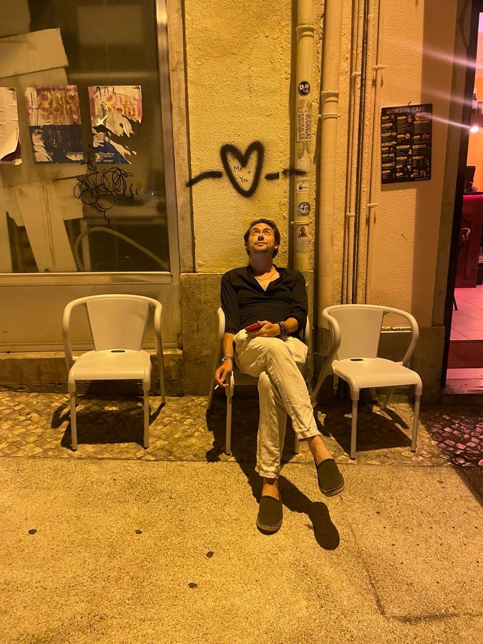 A young man relaxes in a chair at night, gazing at the sky and a heart on the wall.