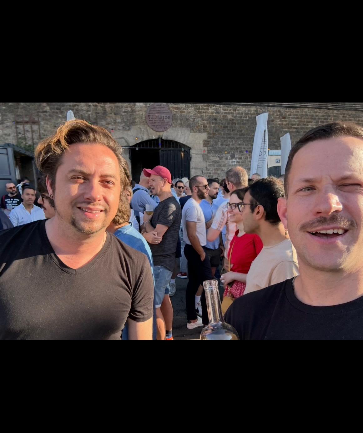 Smiling friends pose together with drinks at a festive outdoor event in a bustling crowd.
