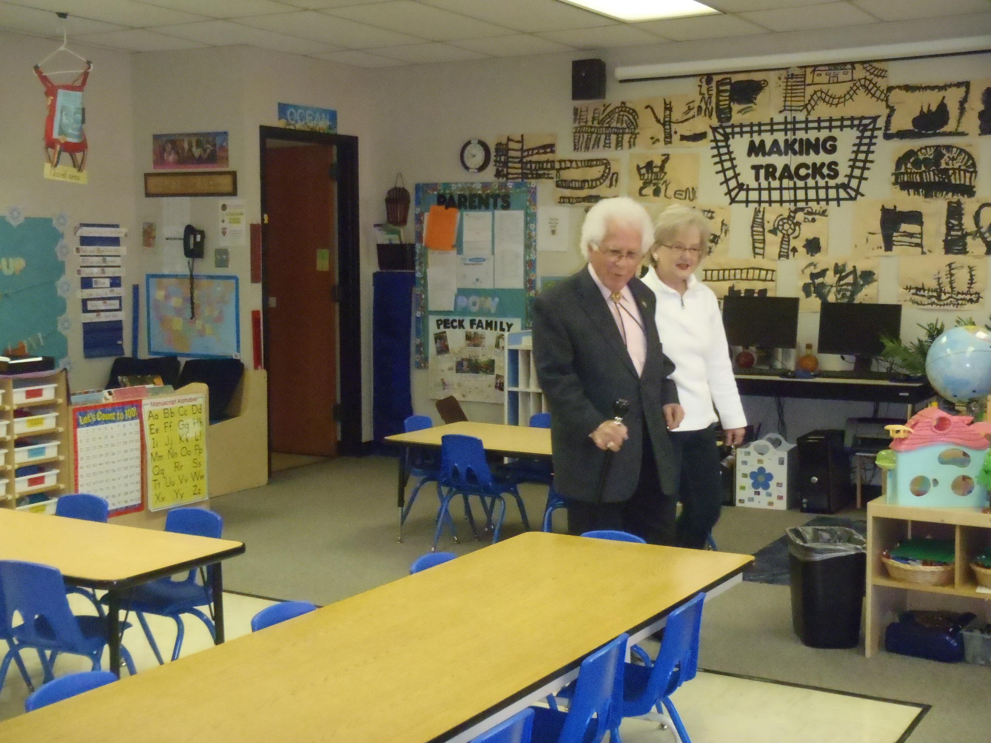Elderly couple walks through a colorful classroom filled with desks and educational materials.