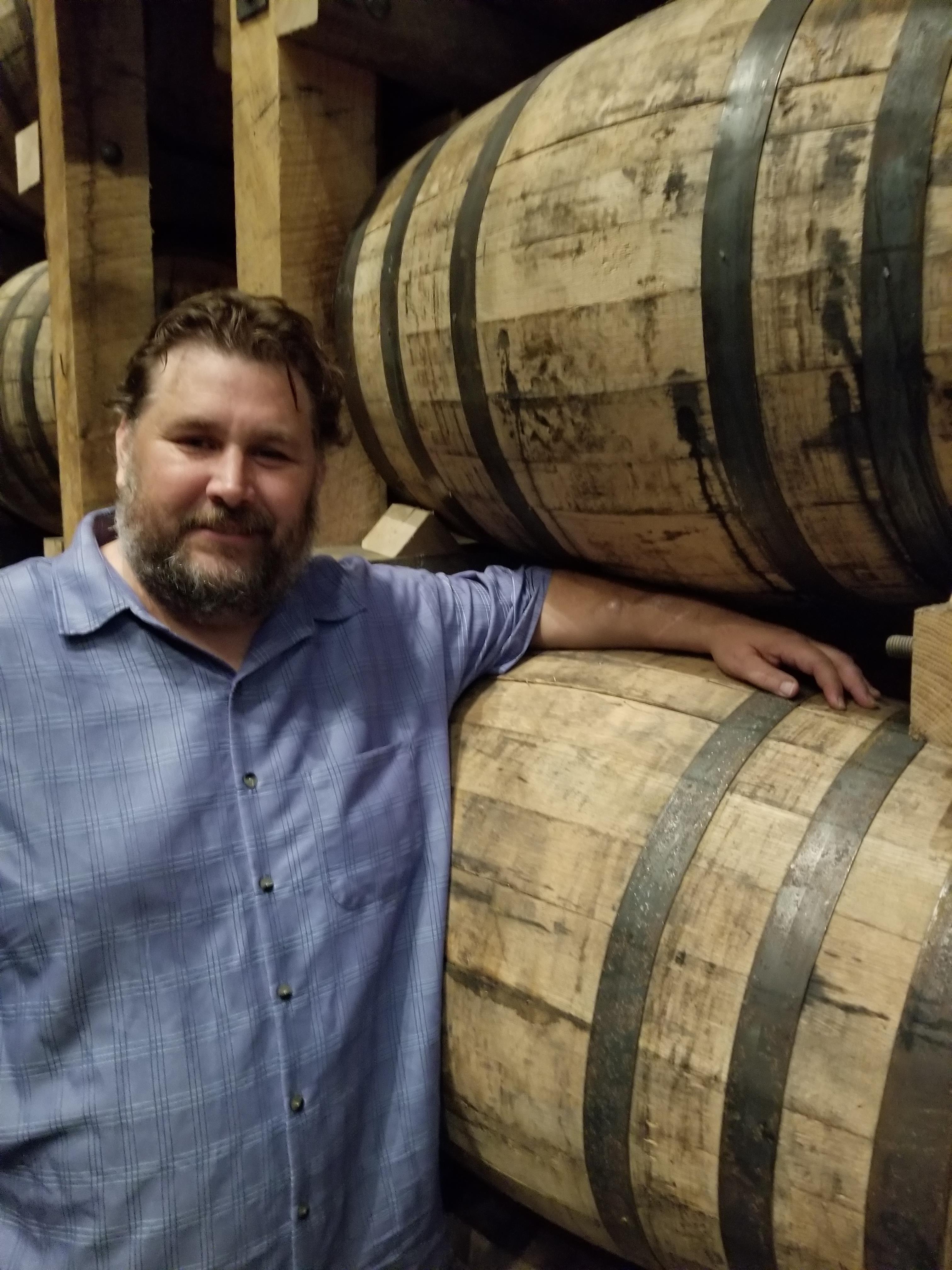 A man stands next to large wooden barrels while enjoying a tasting event at a distillery.