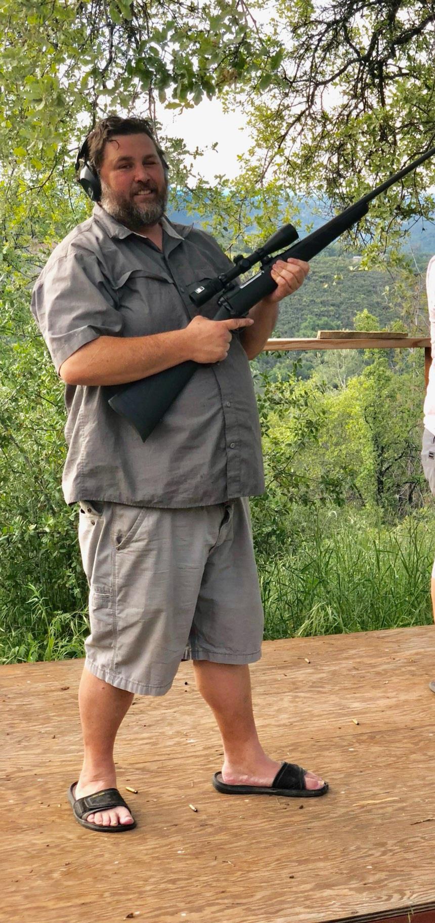 A confident man in shorts stands with a rifle at a shooting range in a green area.