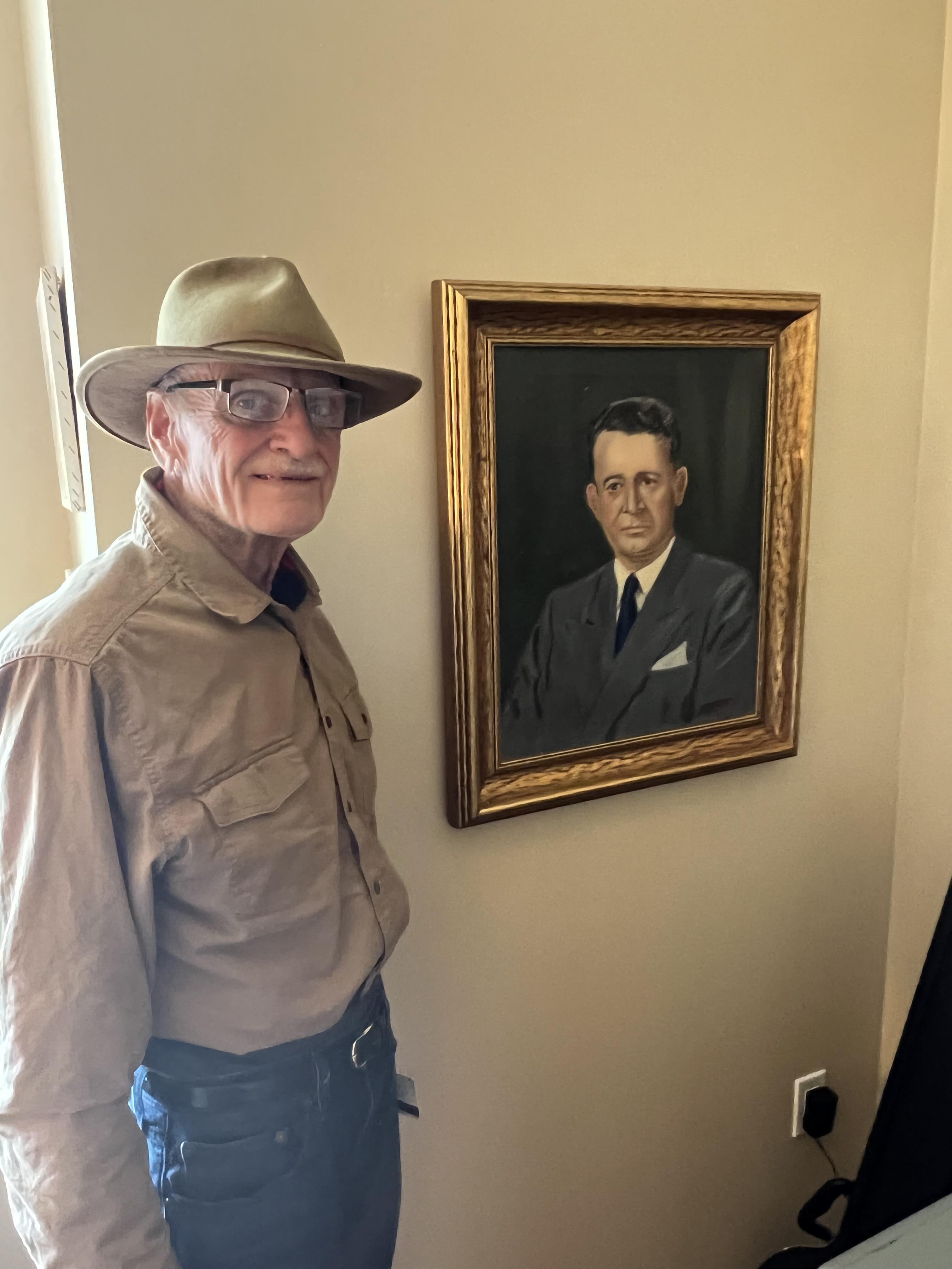 An older gentleman with glasses and a hat poses next to a painted portrait on the wall.