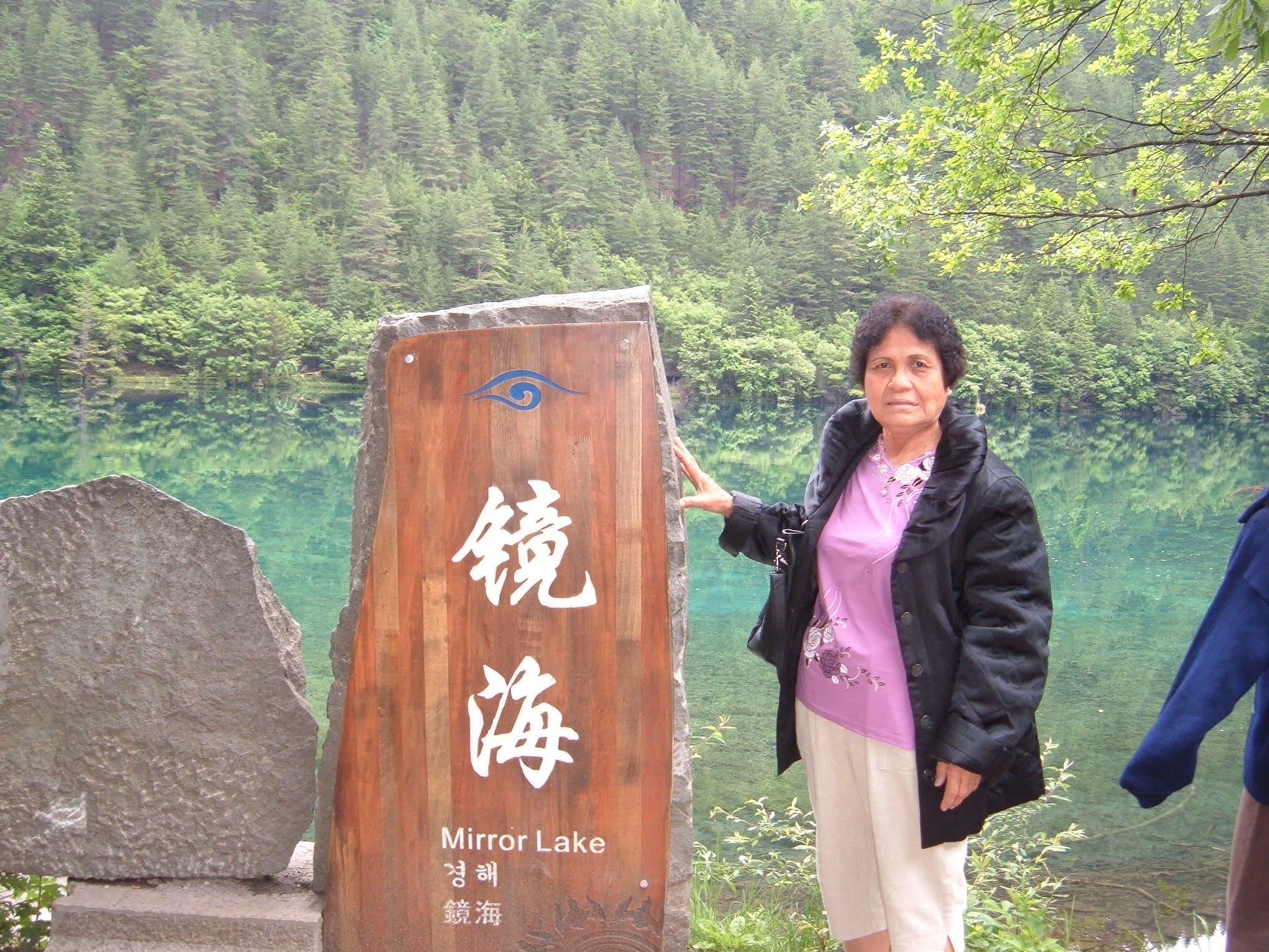 A woman stands beside a wooden sign at Mirror Lake, surrounded by lush greenery and clear water.