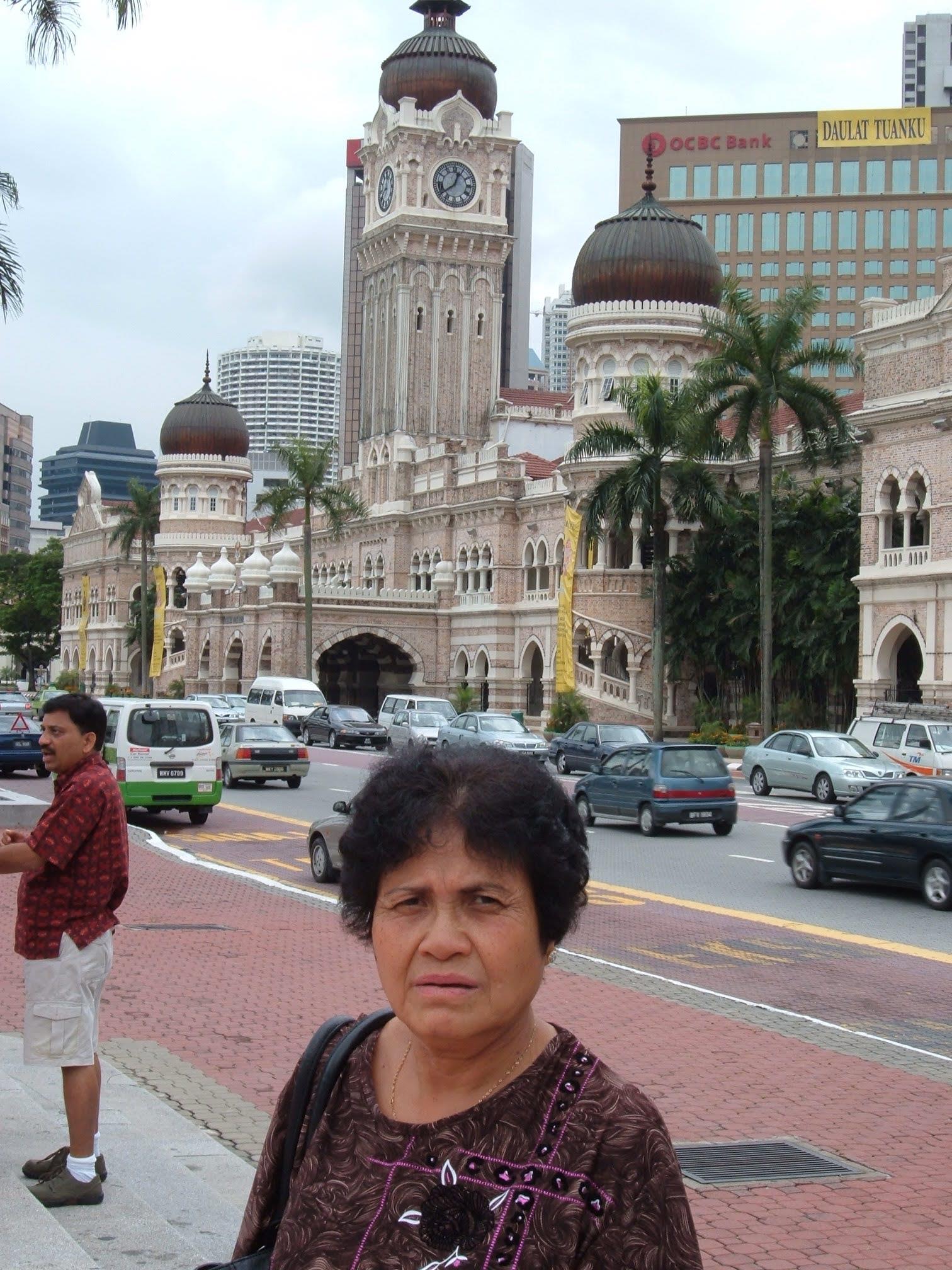 A woman poses for a photo near a classic clock tower surrounded by cars and trees.