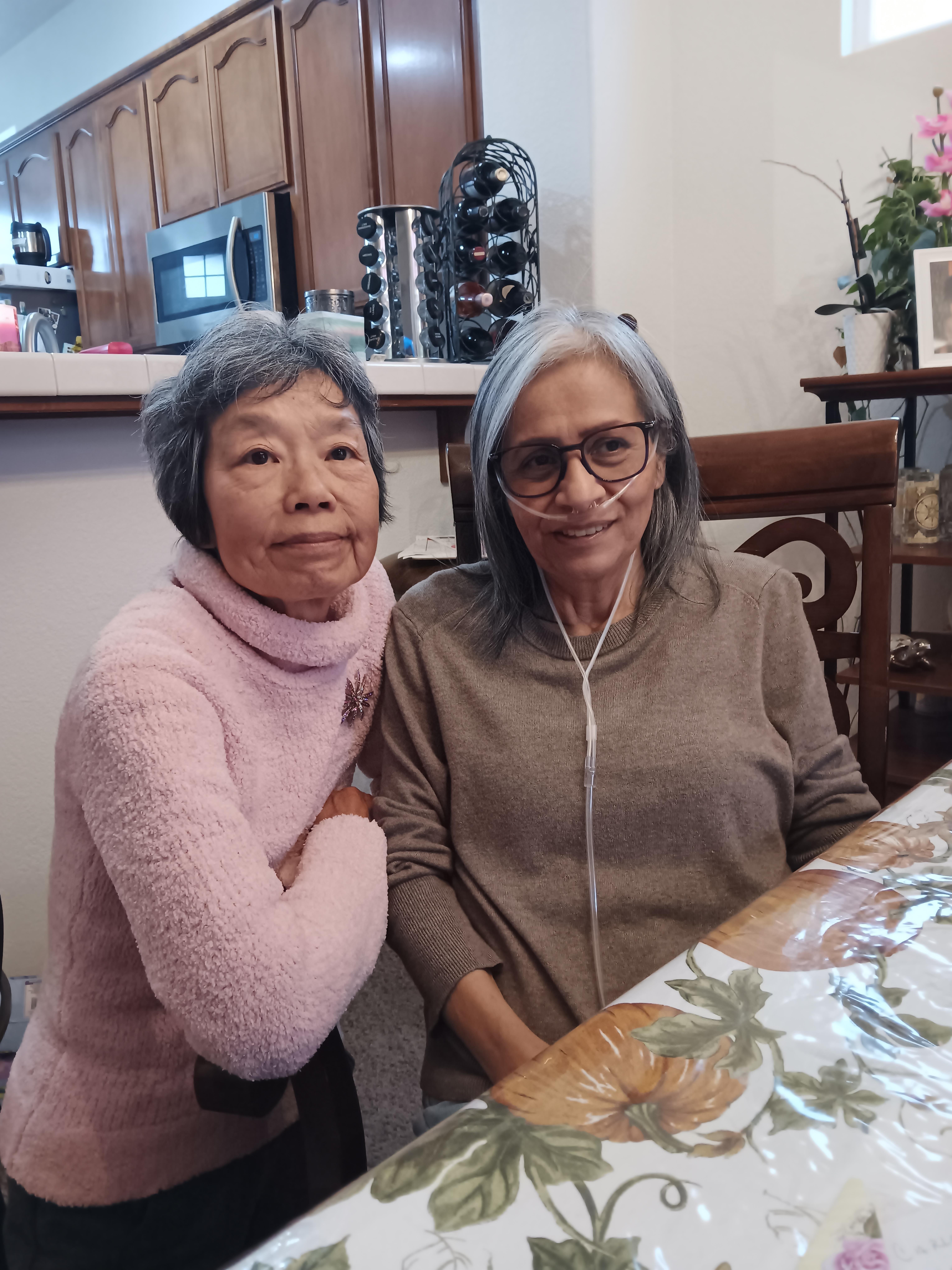 Two elderly women are enjoying each other's company at a kitchen table, smiling happily.
