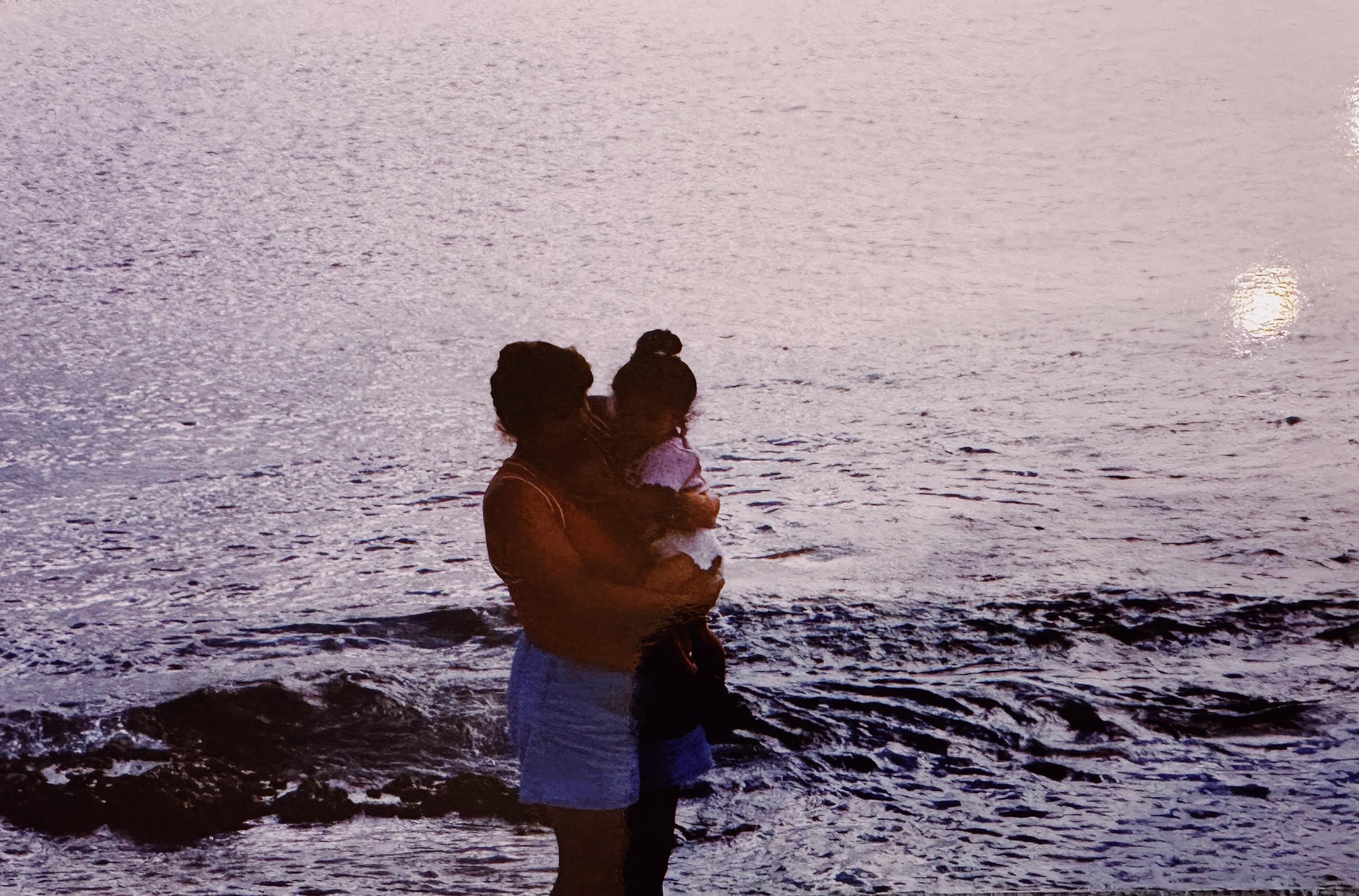 A mother holds her child near the water's edge as waves gently lap the shore at sunset.
