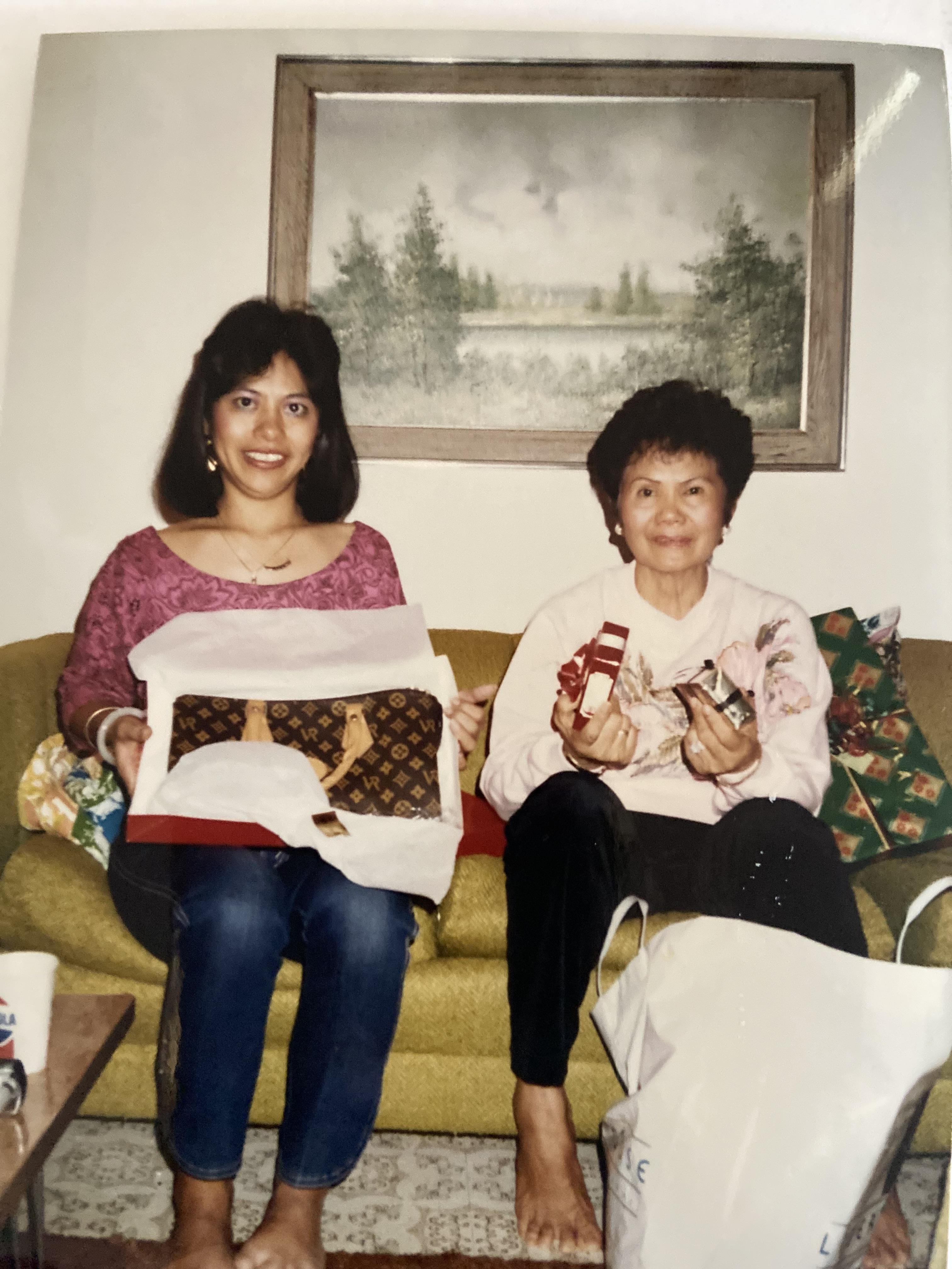 Two women share a joyful moment opening gifts on a cozy couch in a living room.