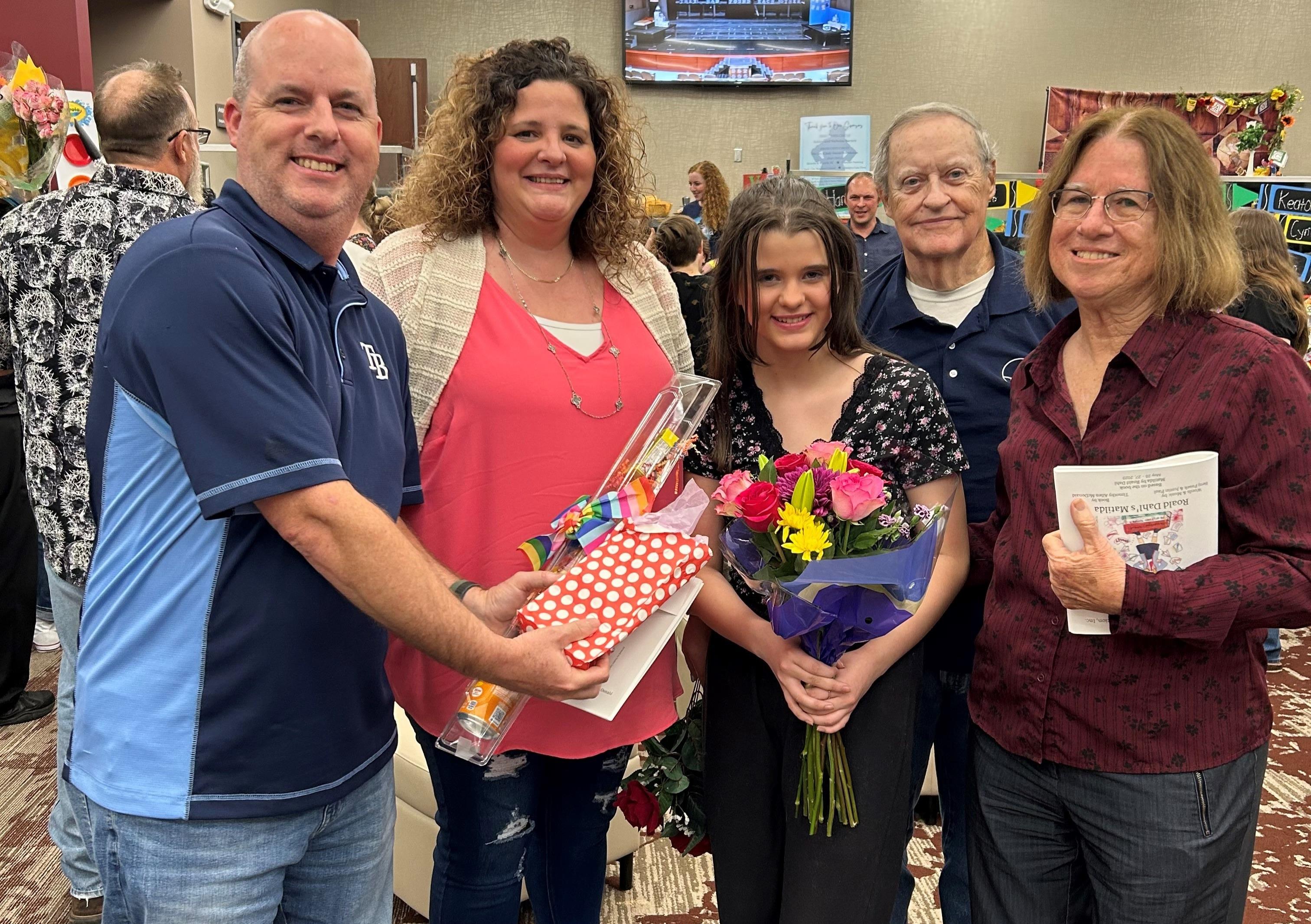 Friends and family gather to celebrate a young girl receiving flowers and gifts at an event.