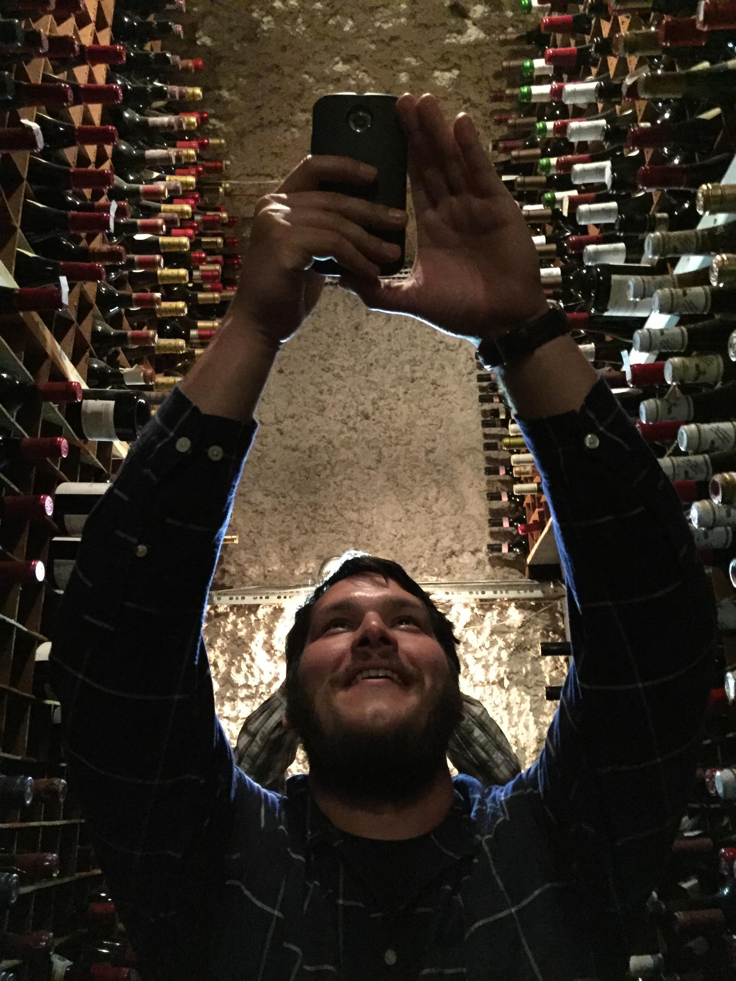 Man smiles while taking a selfie in a well-stocked wine cellar at a local winery.