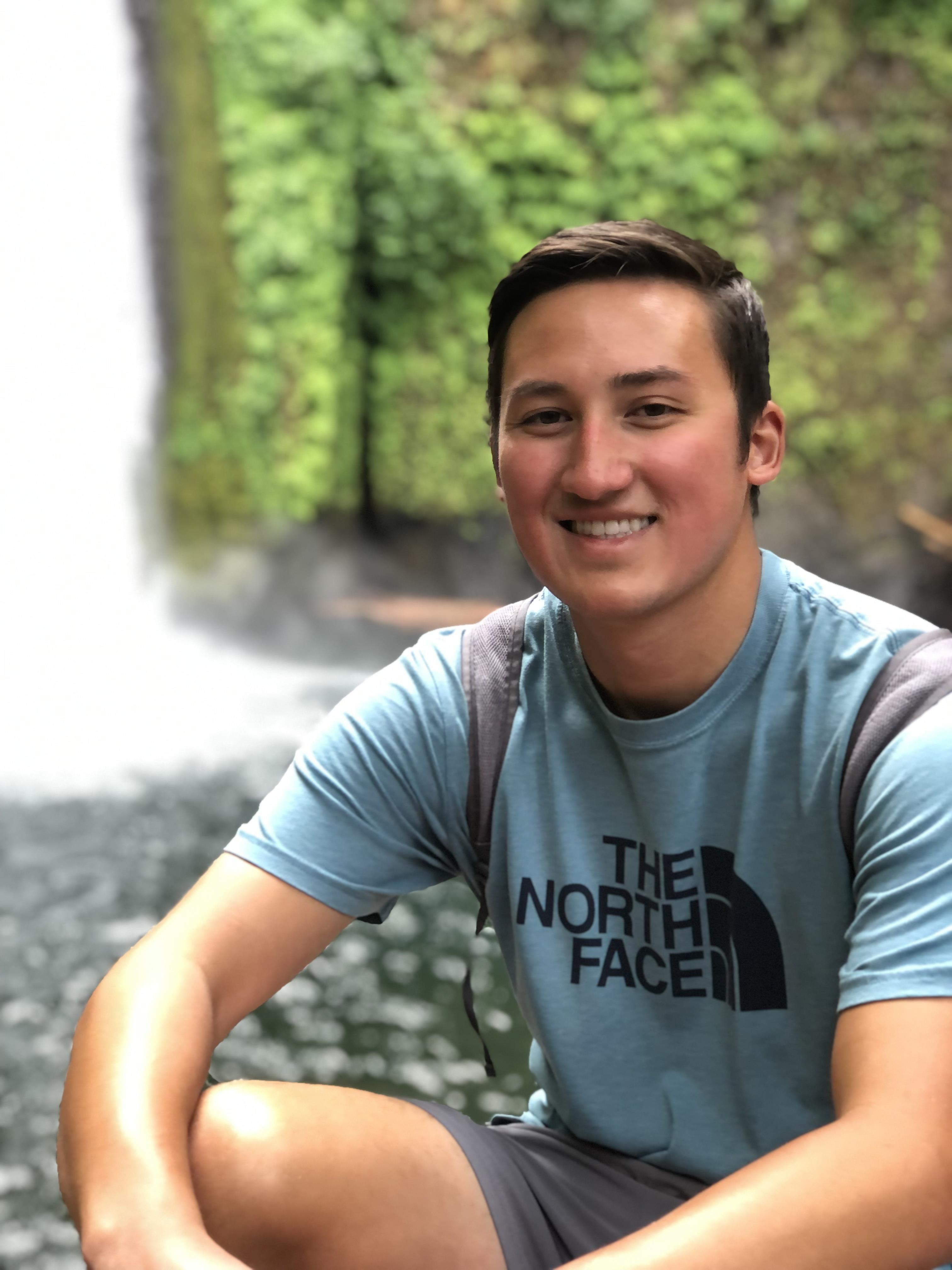 A smiling young man relaxes by a tranquil waterfall surrounded by lush greenery.