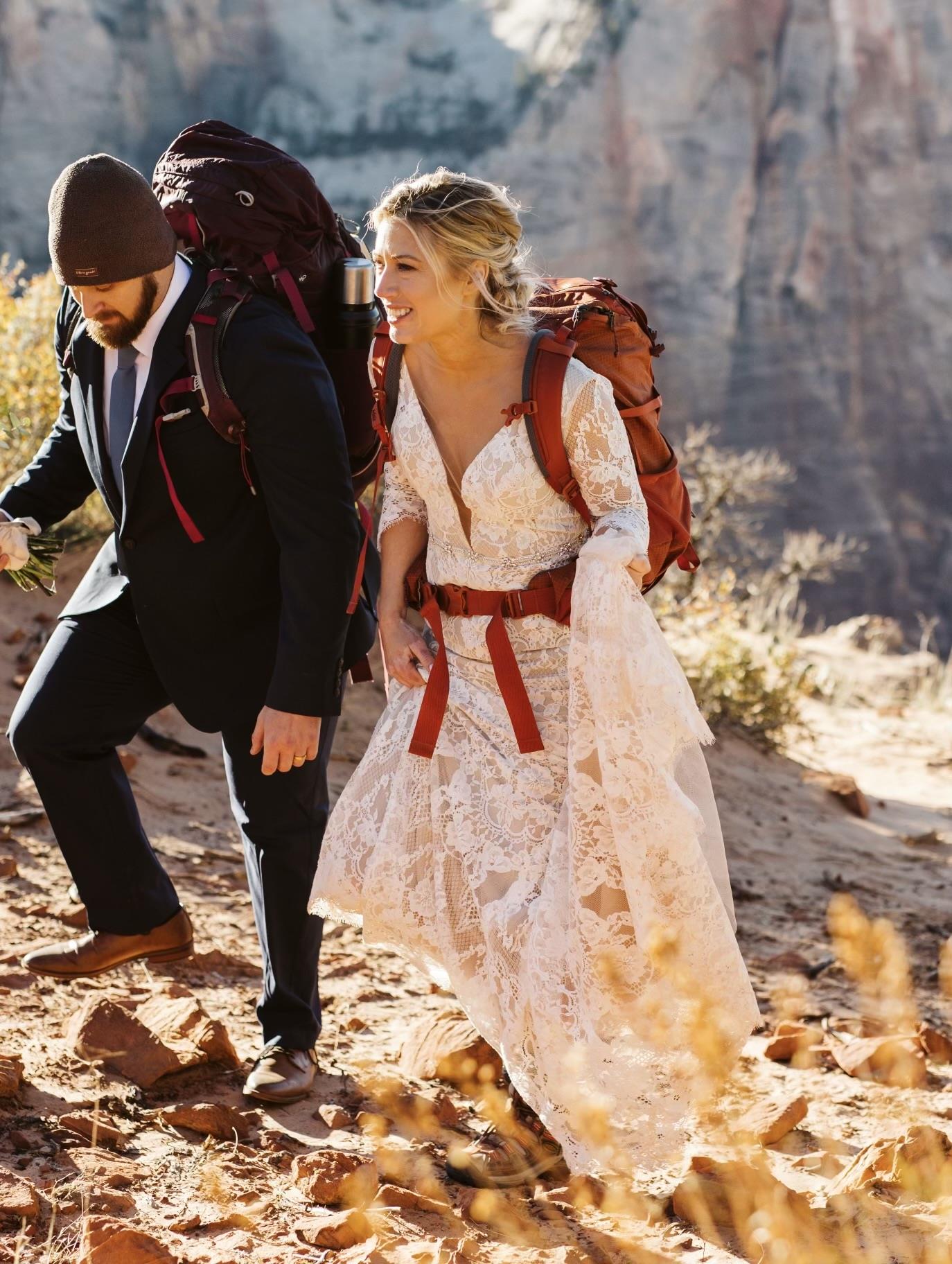 A couple dressed in wedding attire and hiking gear climbs a rocky trail in a national park.
