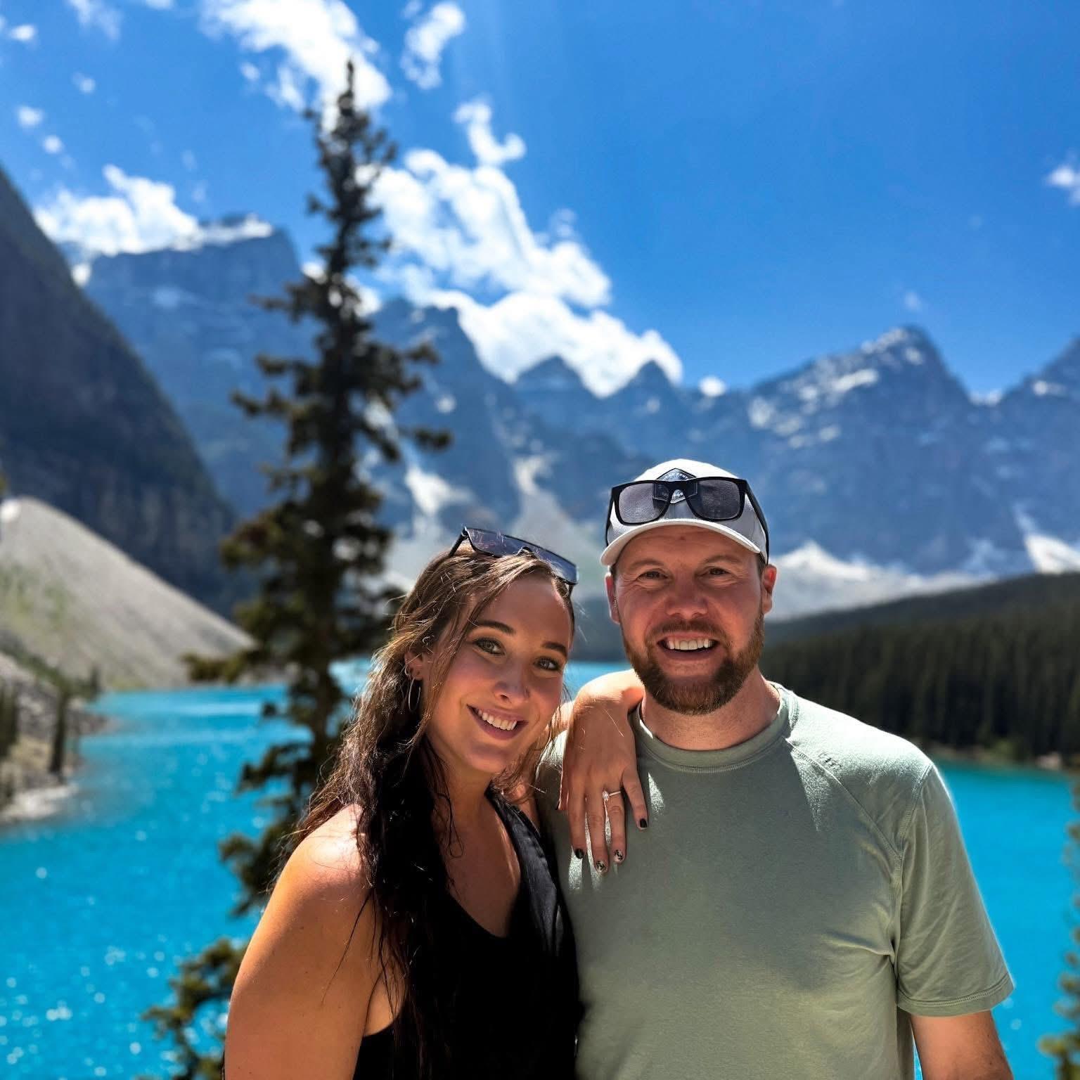 A couple smiles with the stunning turquoise lake and mountain backdrop on a bright day.