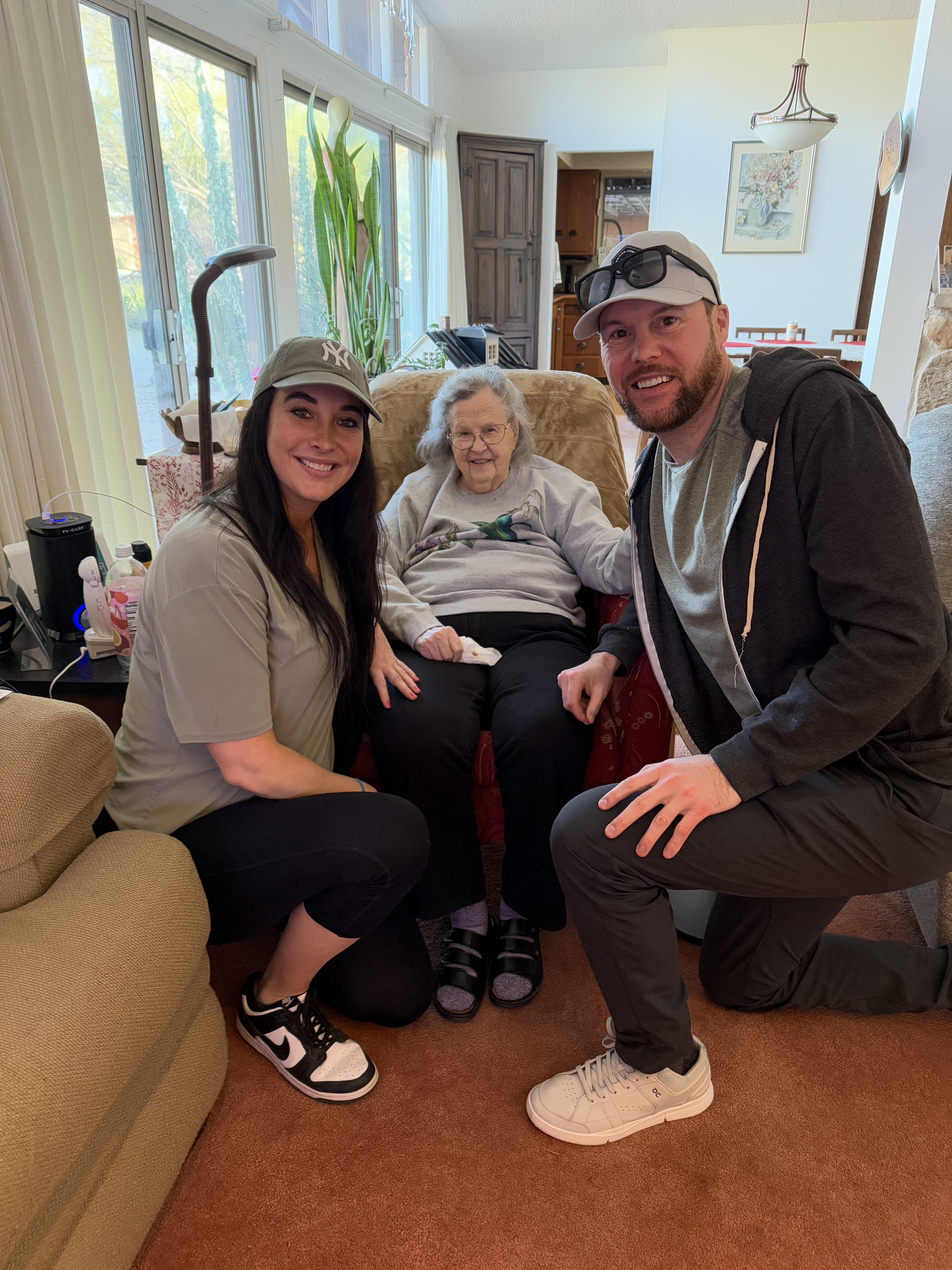 Two adults visit an elderly woman, sharing smiles in her cozy living room.