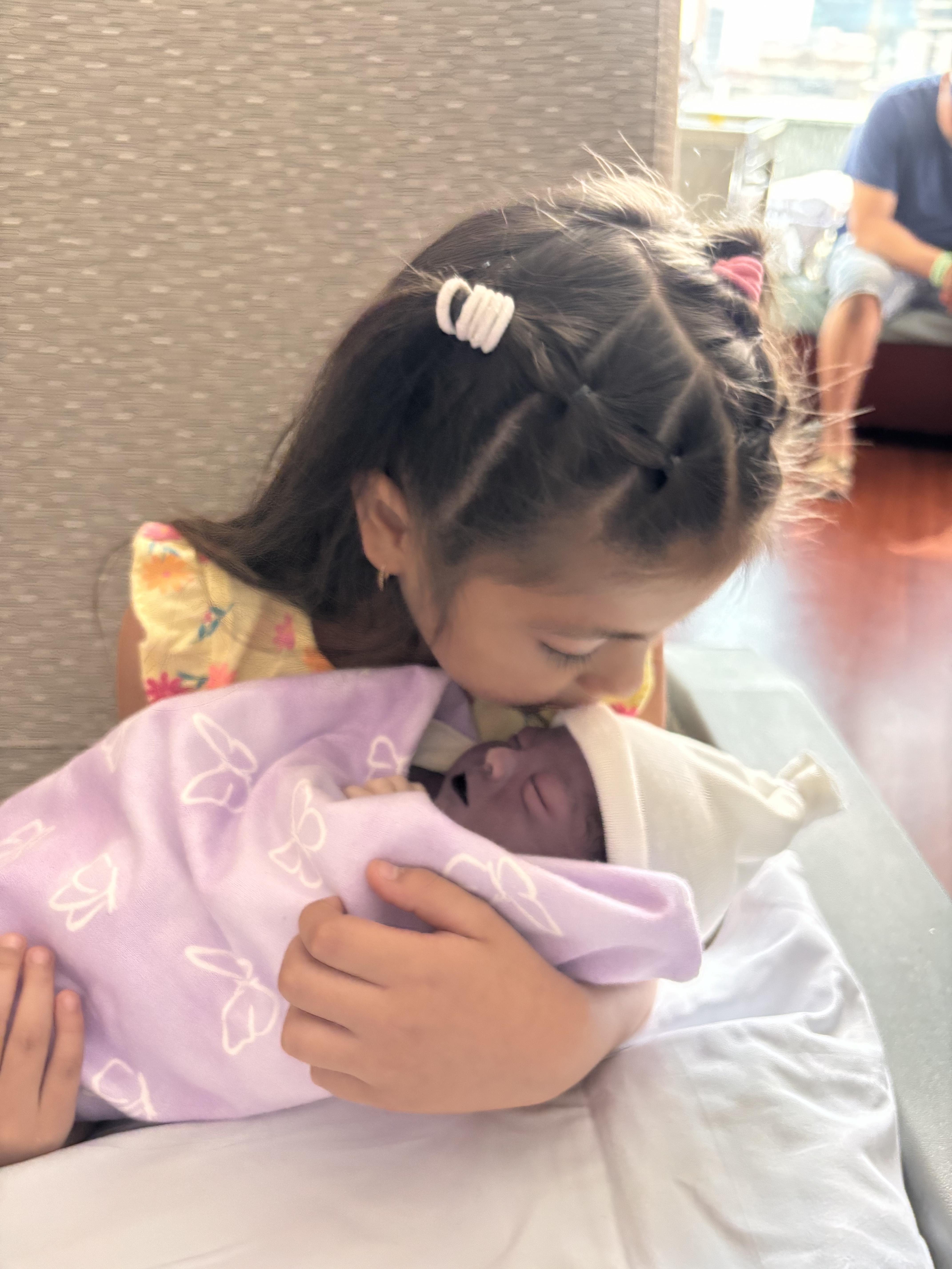 A girl embraces her newborn sibling in a cozy setting, sharing a tender kiss.