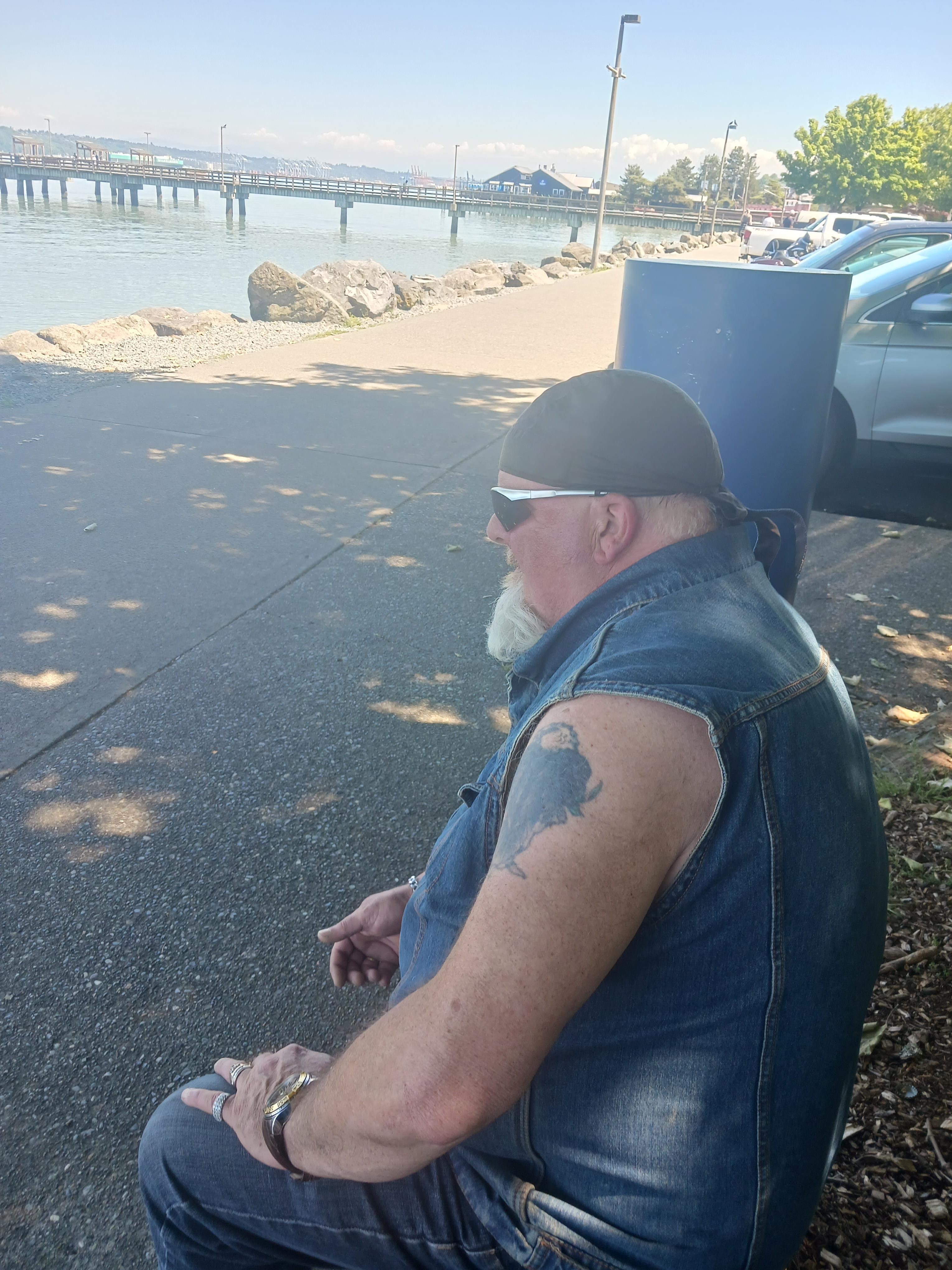 A bearded man in a denim vest relaxes by the water, smoking a cigarette and enjoying the view.