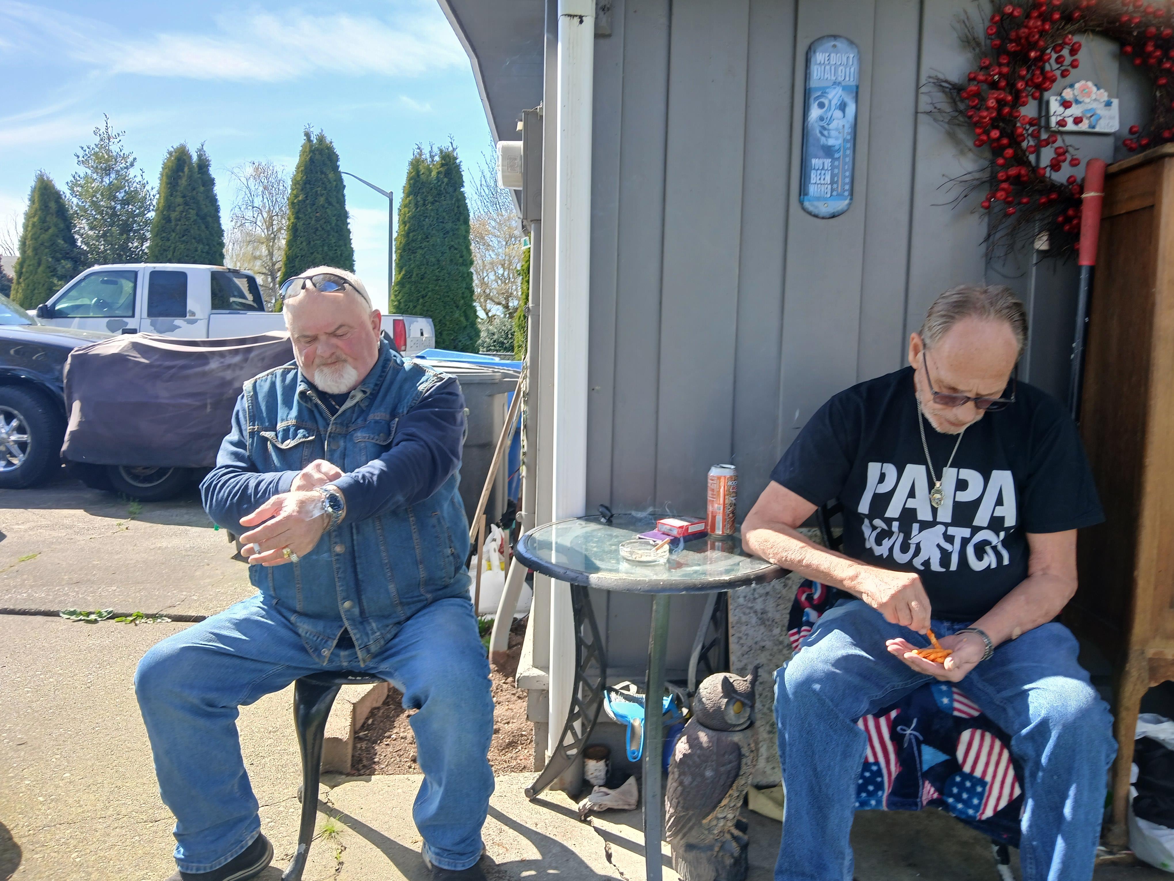 Two men sit outside a house, one is wearing a denim jacket, and both are enjoying their drinks.