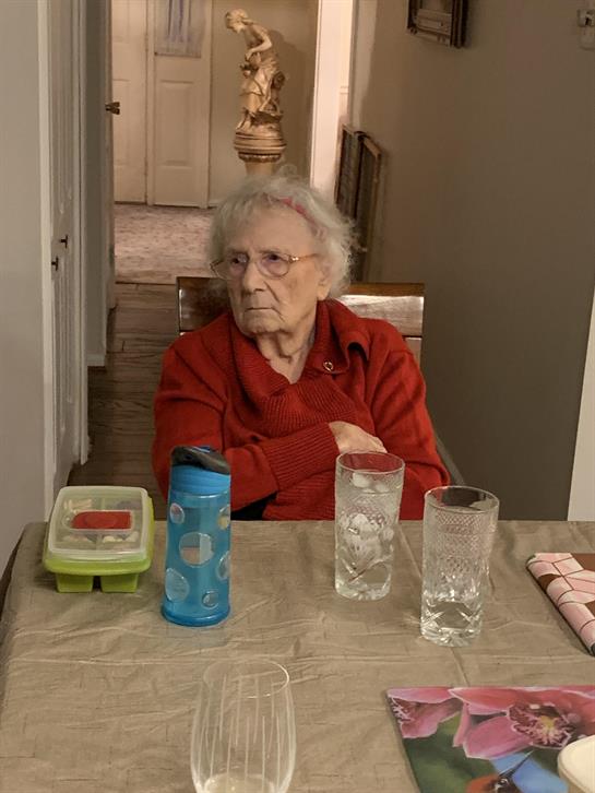 An old woman sitting at a table with glasses and a thermos