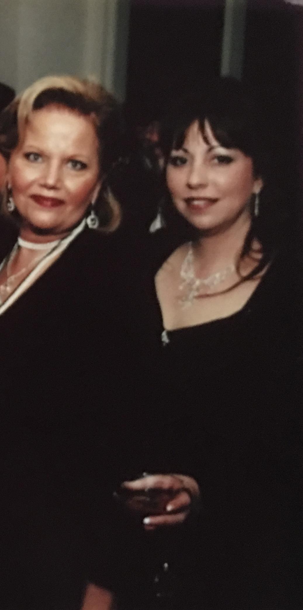 Two women socialize at a formal event, wearing elegant outfits and adorned with jewelry.