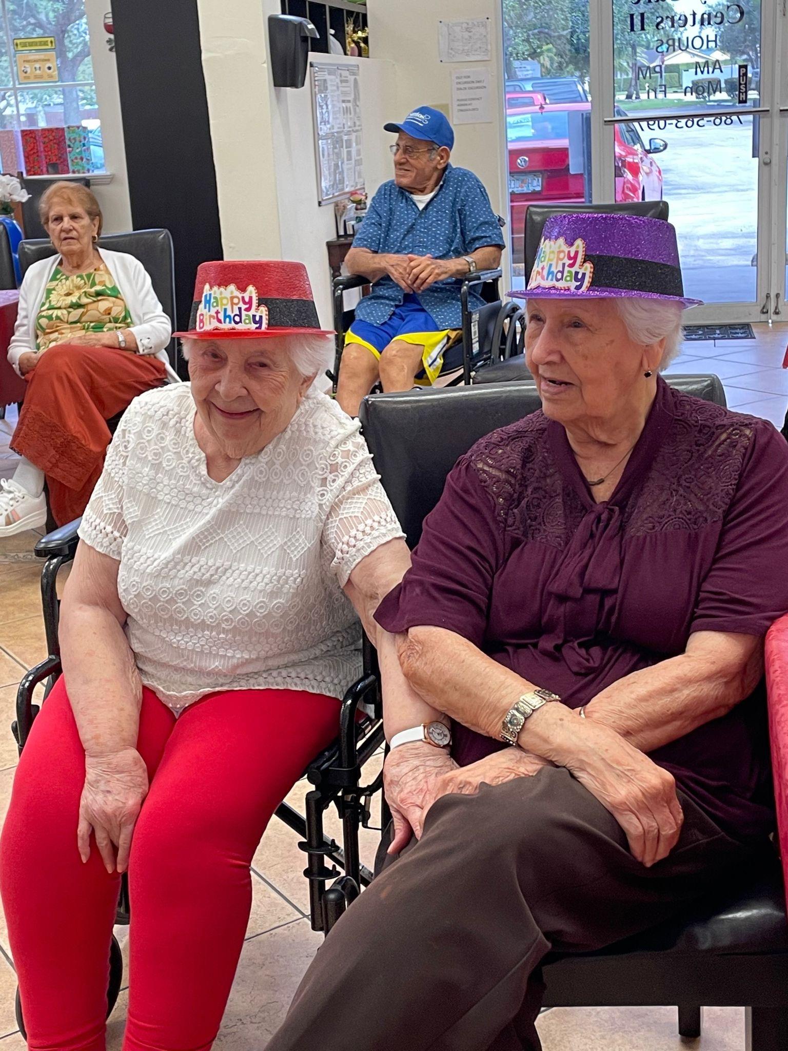 Two elderly women in colorful hats enjoy the festive atmosphere at a care center celebration.