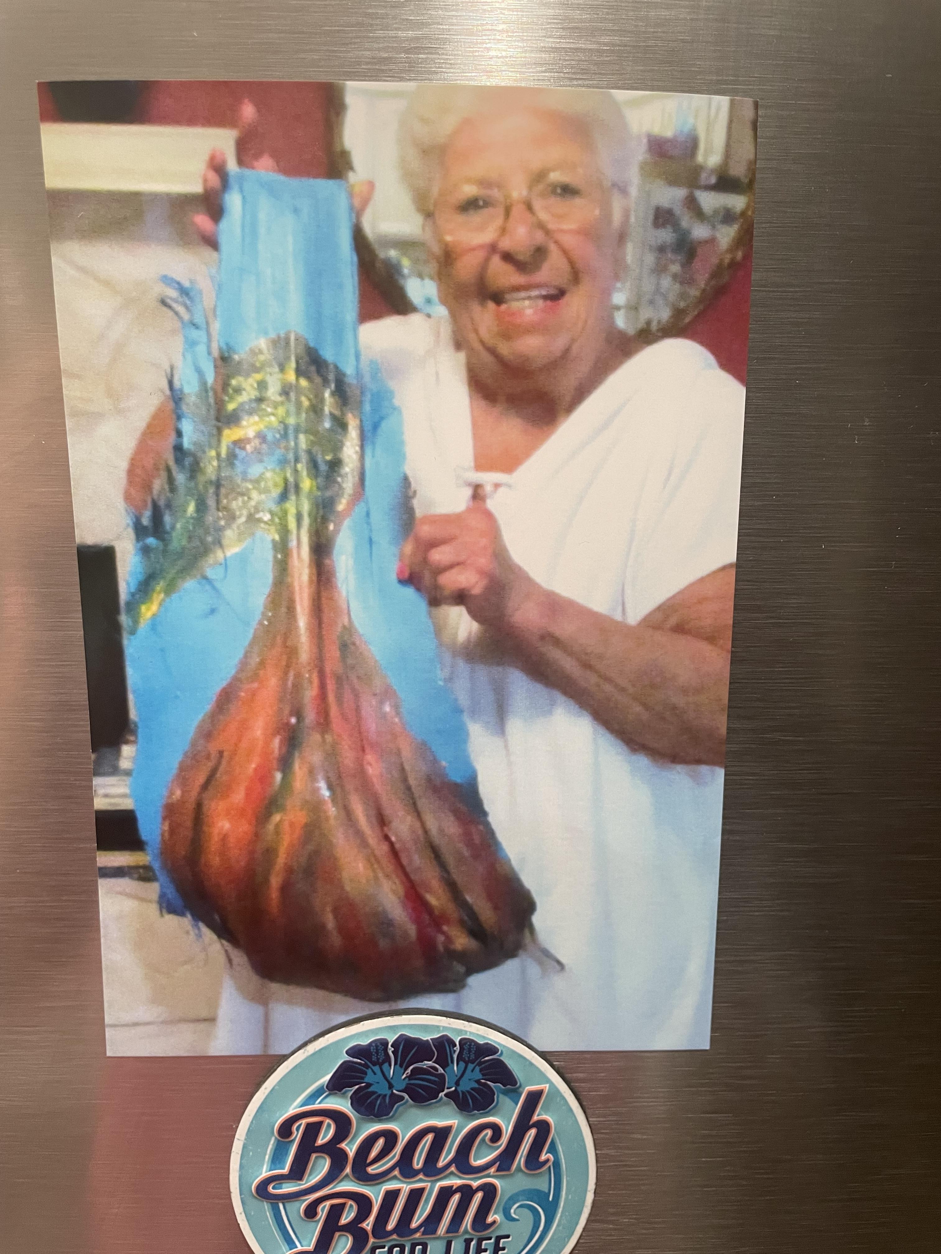 Elderly woman smiles while holding colorful handcrafted art in a cozy kitchen.